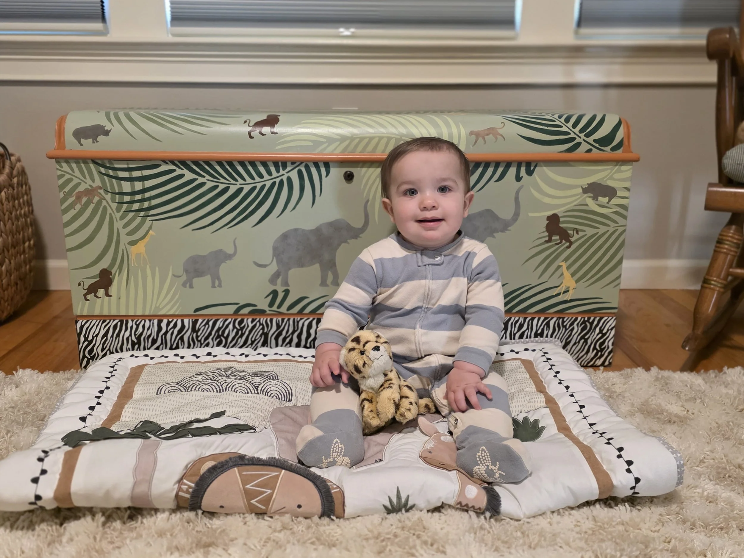 A young child sitting on a patterned quilt on a fluffy rug, holding a plush tiger toy, with a toy drum and decorative wall covering featuring jungle animals in the background.