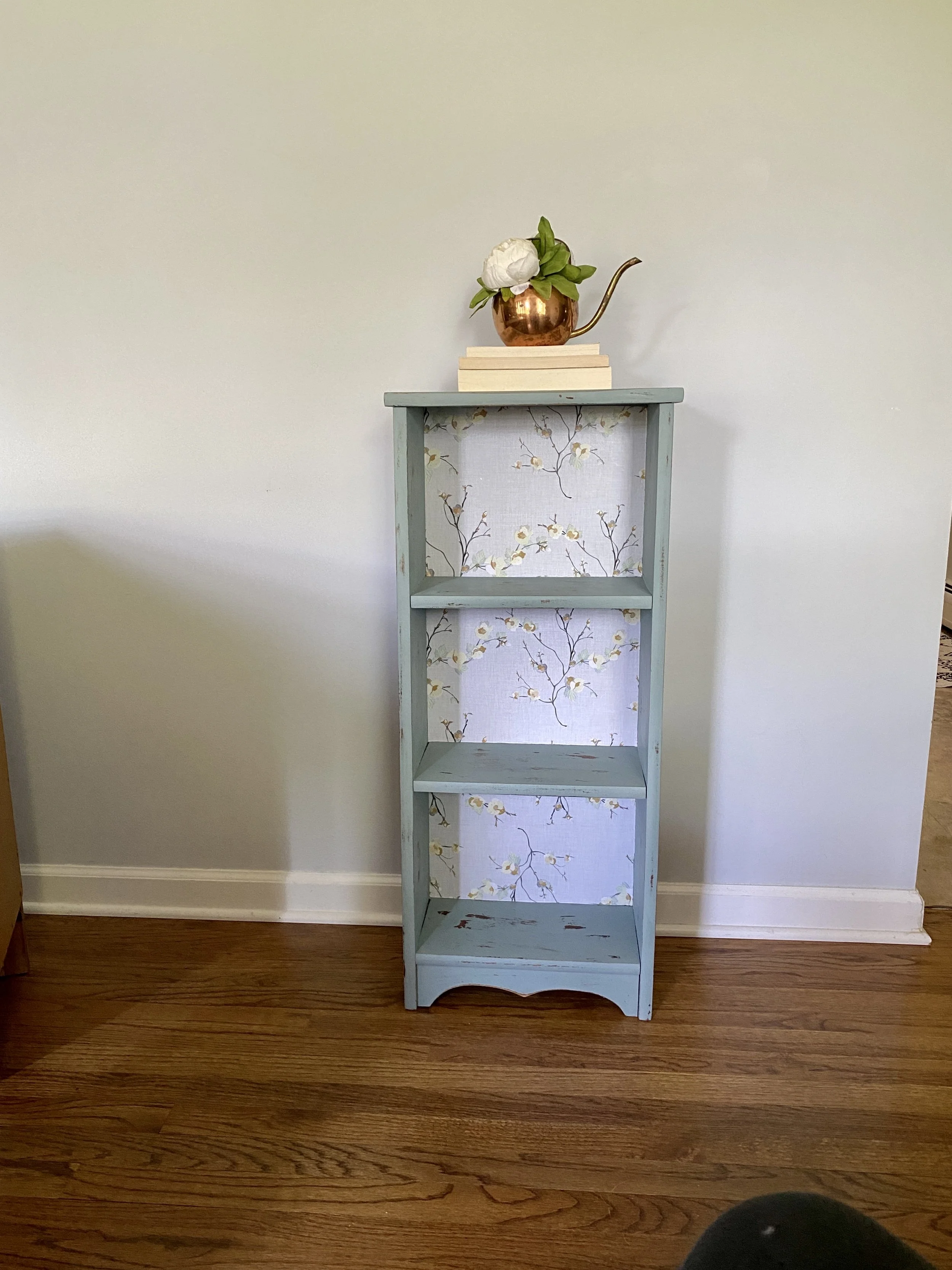 A tall, narrow, light blue wooden shelf with four shelves, the second shelf from the top has a floral-patterned backing. On top of the shelf, there is a stack of books with a copper watering can containing a white peony flower and green leaves placed