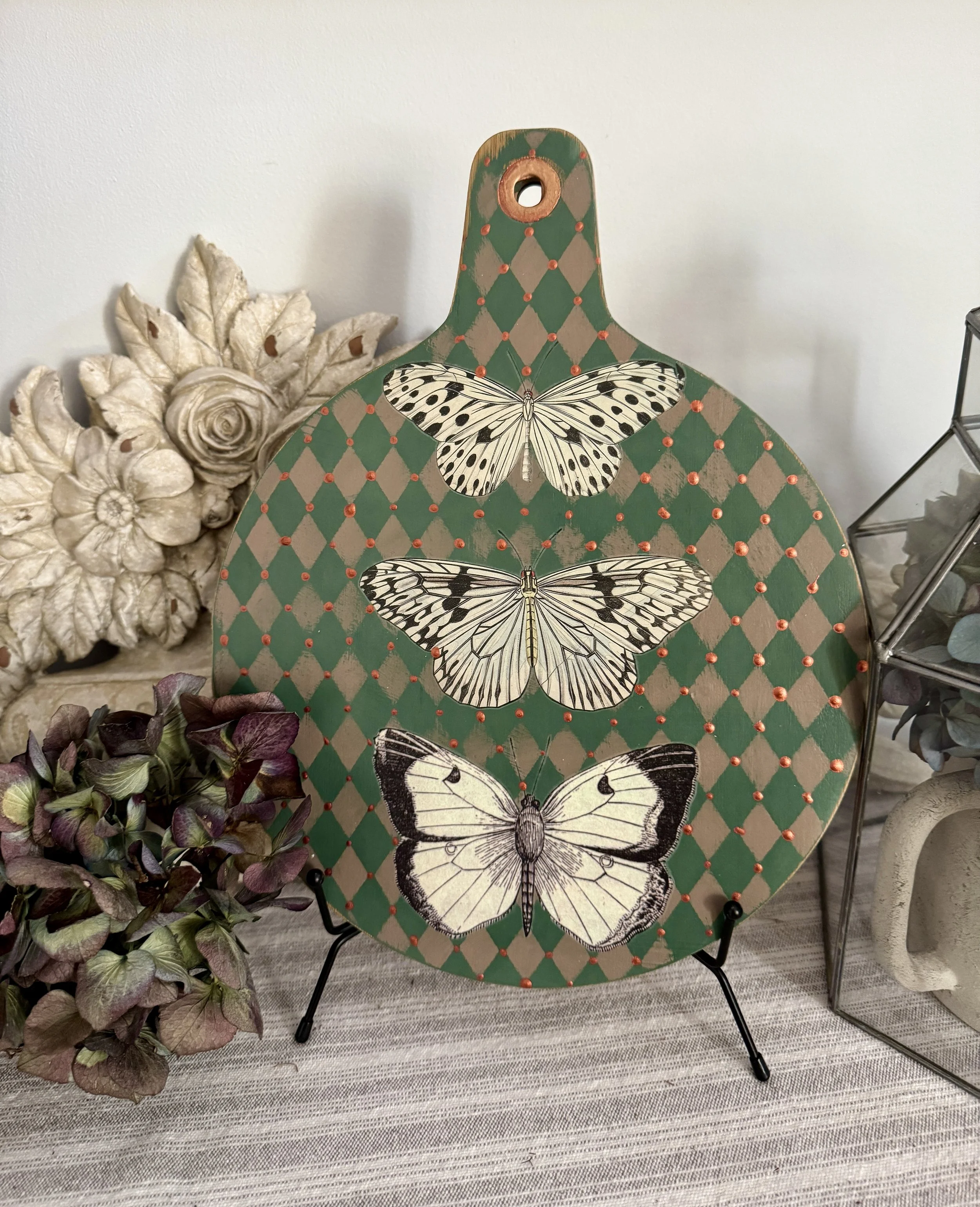 Decorative platter with three black and white butterfly illustrations, surrounded by dried flowers including a cream-colored rose and purple-green hydrangea, placed on a textured beige and gray striped rug.
