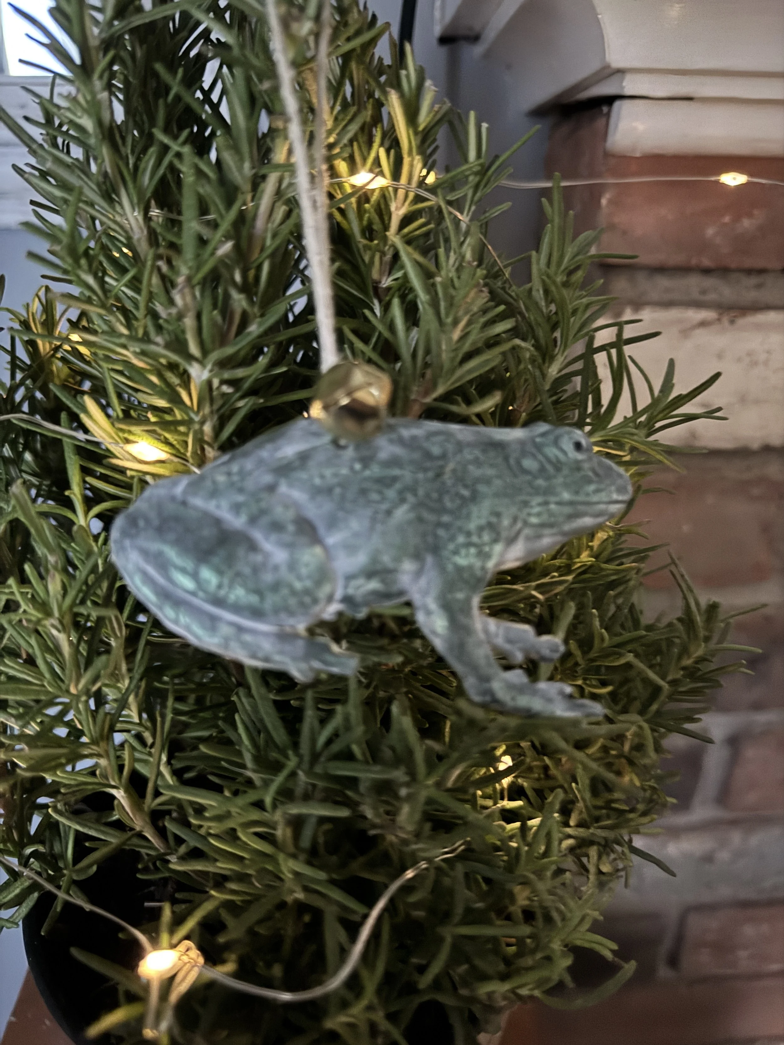 Close-up of a small artificial frog sculpture resting on a lush green plant adorned with a string of small warm white fairy lights, with a wooden and brick background.