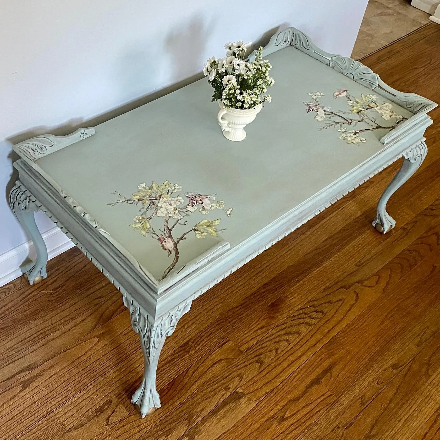A vintage style coffee table with ornate carved legs and a painted floral design on the surface, topped with a white vase of white daisies.