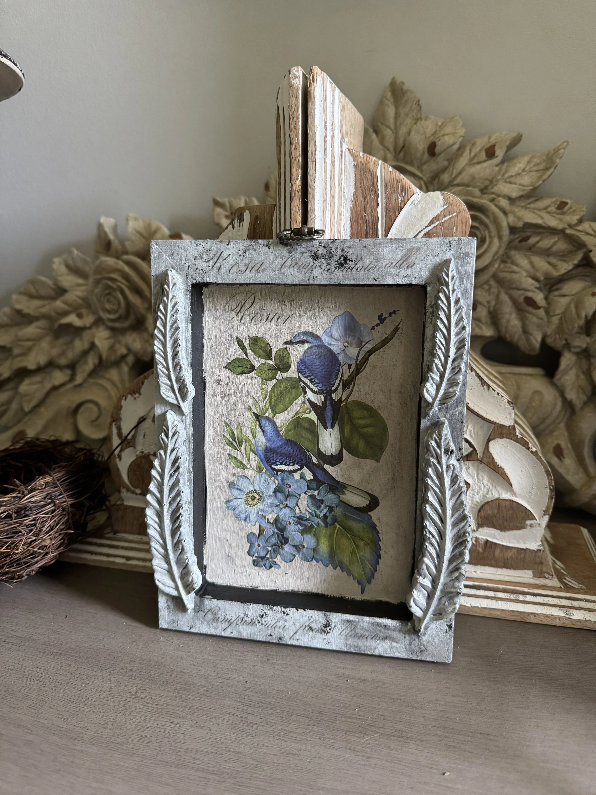 Decorative picture frame with a floral print of blue birds and flowers, surrounded by a white ornate border, on a wooden surface with carved wood leaves in the background.