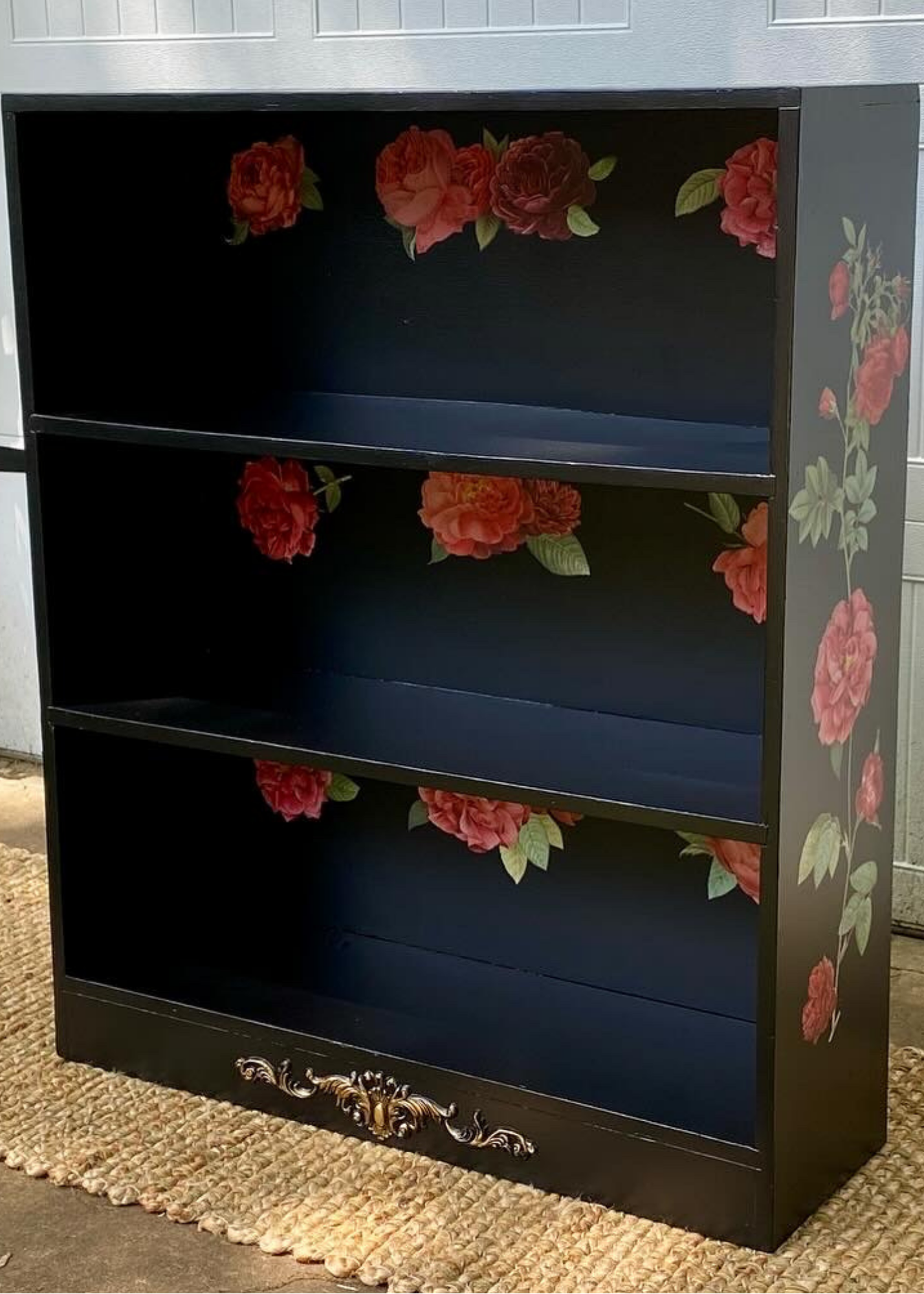Black bookshelf with floral design on the sides and bottom, featuring red and pink flowers with green leaves painted on the sides and inside shelves, placed on a woven rug outdoors.