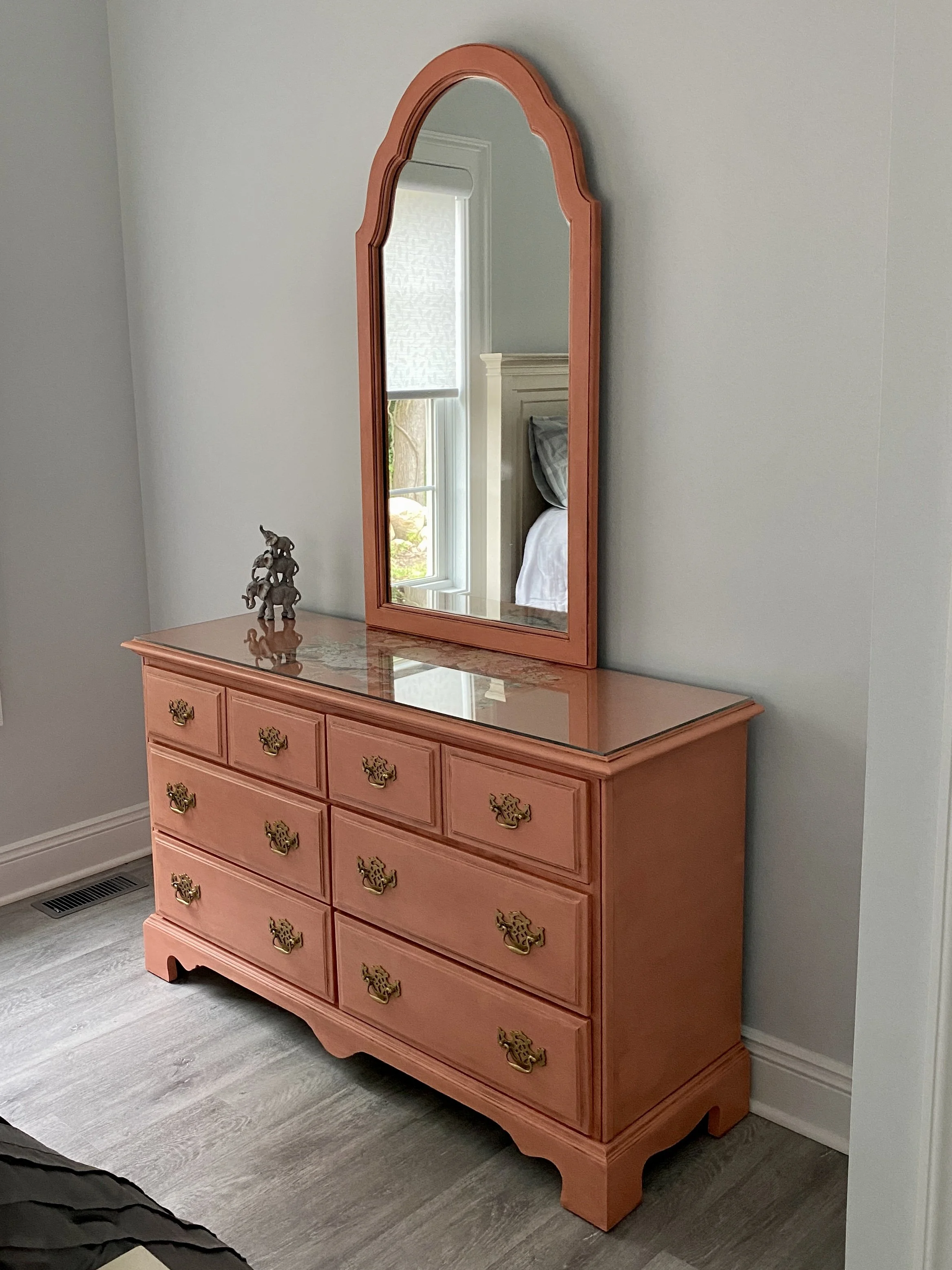 A pink wooden dresser with gold handles and a matching mirror. A small decorative figurine of elephants is on top of the dresser.