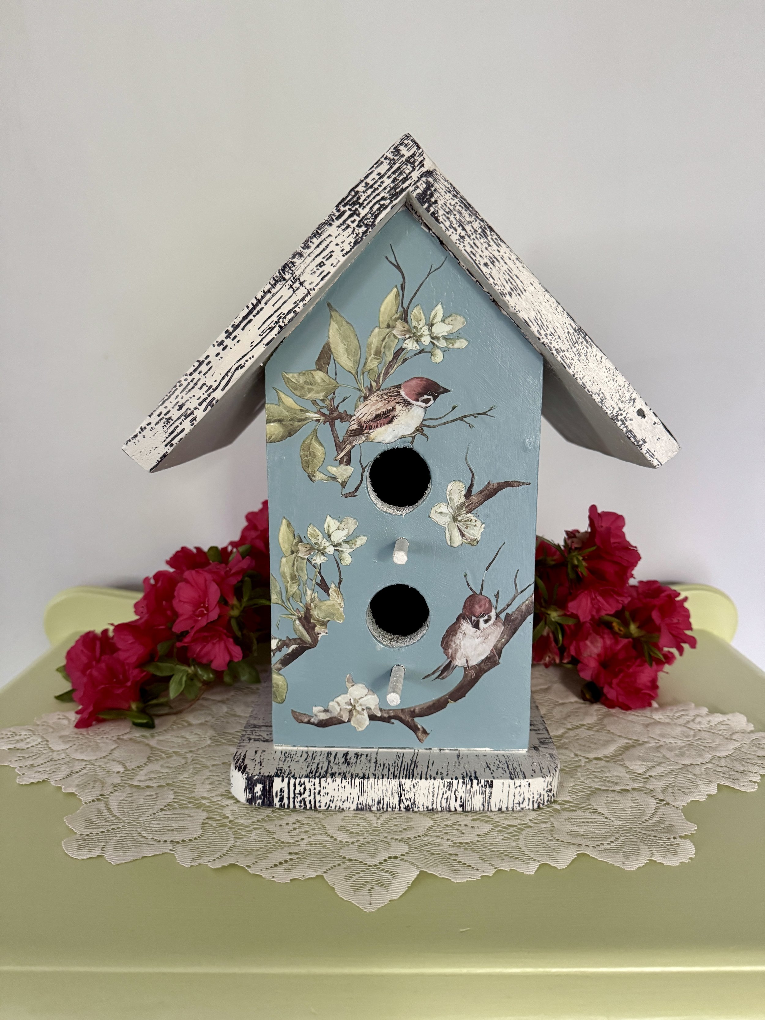 Decorative blue birdhouse with painted birds and blossoms, surrounded by pink flowers on a lace-covered surface.