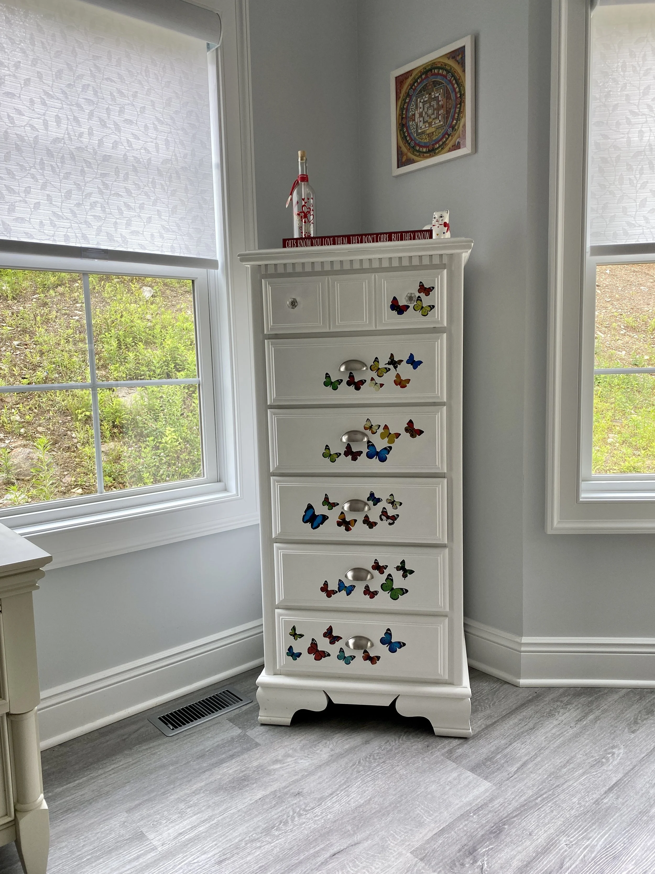 A white dresser with colorful butterfly stickers, situated next to a window with white blinds, in a room with light gray walls. A framed colorful artwork hangs on the wall above the dresser.
