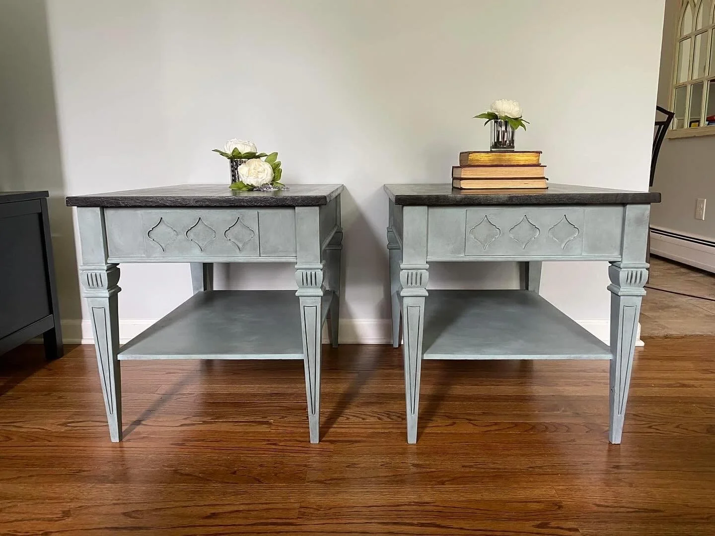 Two matching vintage-style gray side tables with dark wood tops, decorated with vases of white flowers and stacked books, standing on a hardwood floor against a plain wall.