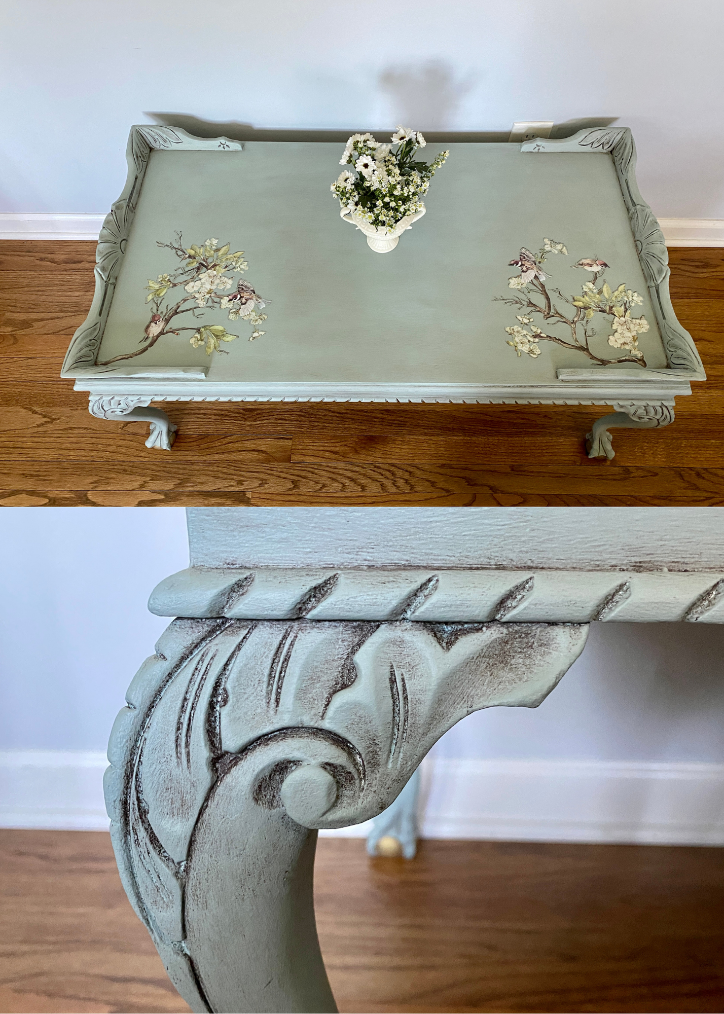 Top view of a vintage style table with ornate carved legs and floral painted design on the surface, a small vase of white flowers in the center, and a wooden floor underneath.