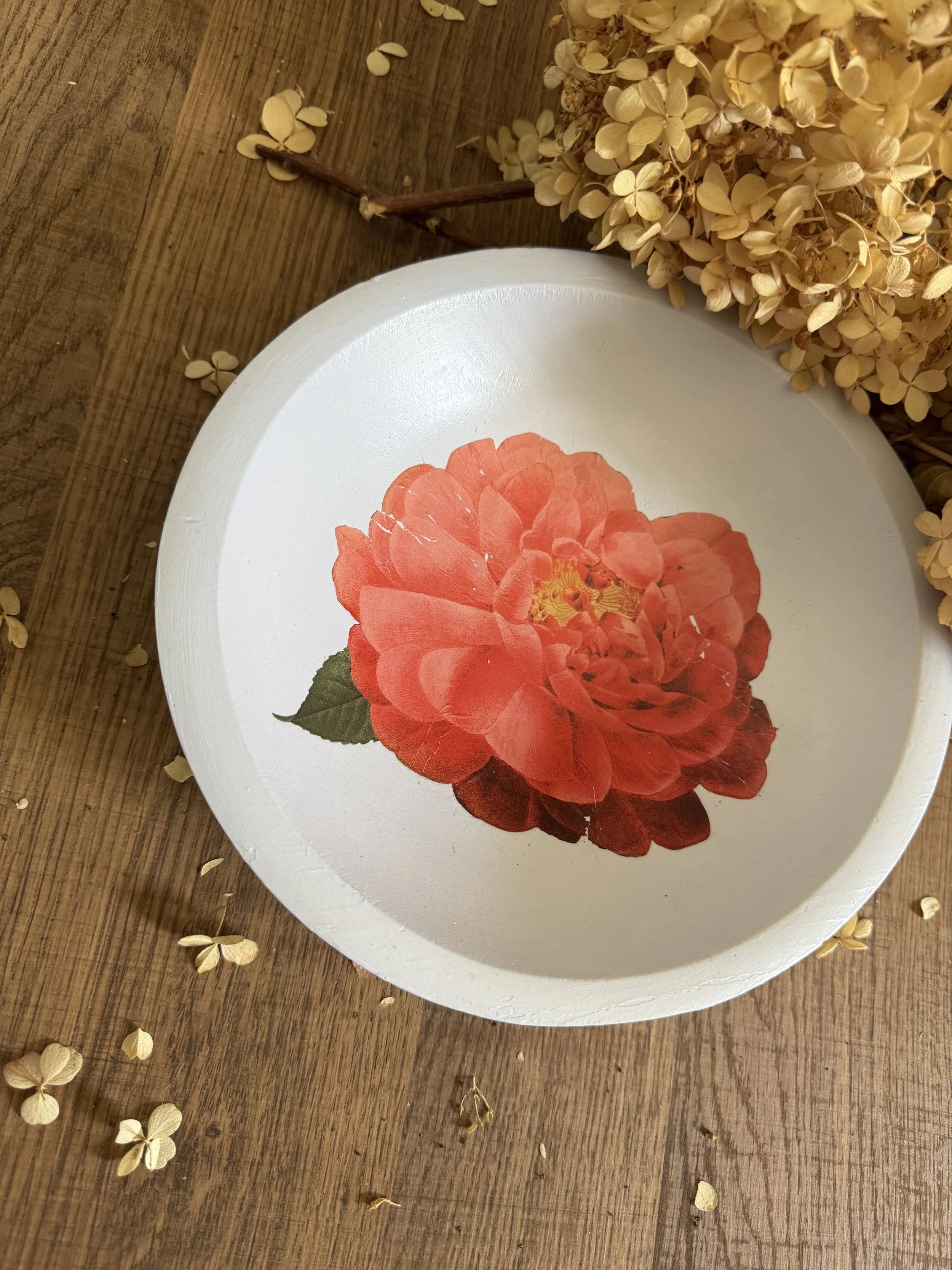 A white circular tray with a large red and orange flower design, placed on a wooden surface with scattered dried flowers.
