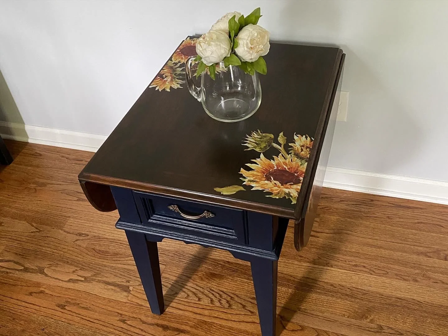A small black side table with a drawer, decorated with sunflower decals on the top, holds a clear glass pitcher with white peonies. The table is on a hardwood floor against a light gray wall.