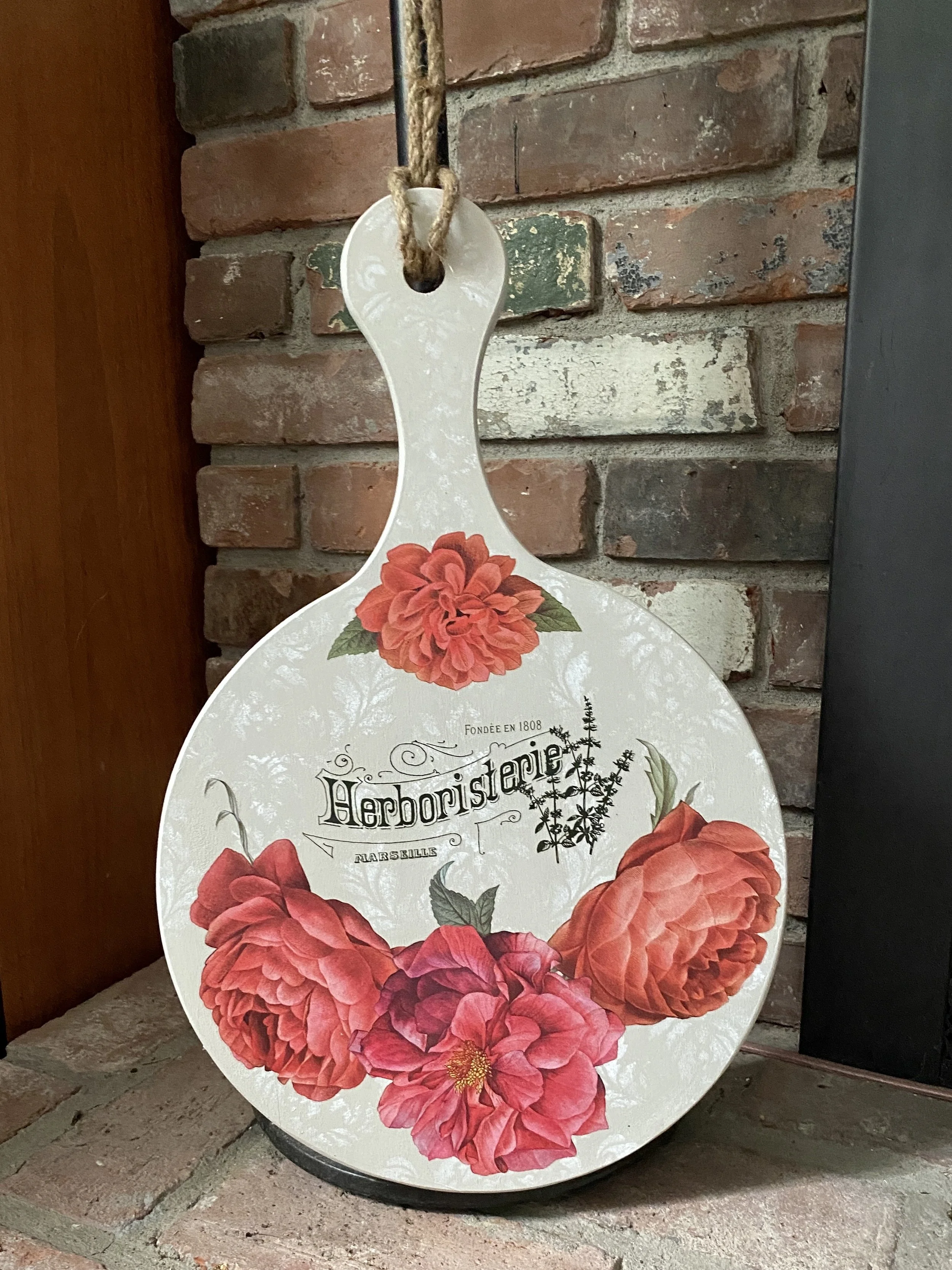 A decorative board with a floral design featuring large red and pink flowers and green leaves, with the word "Herboristerie" and additional text in French, hanging on a brick wall.