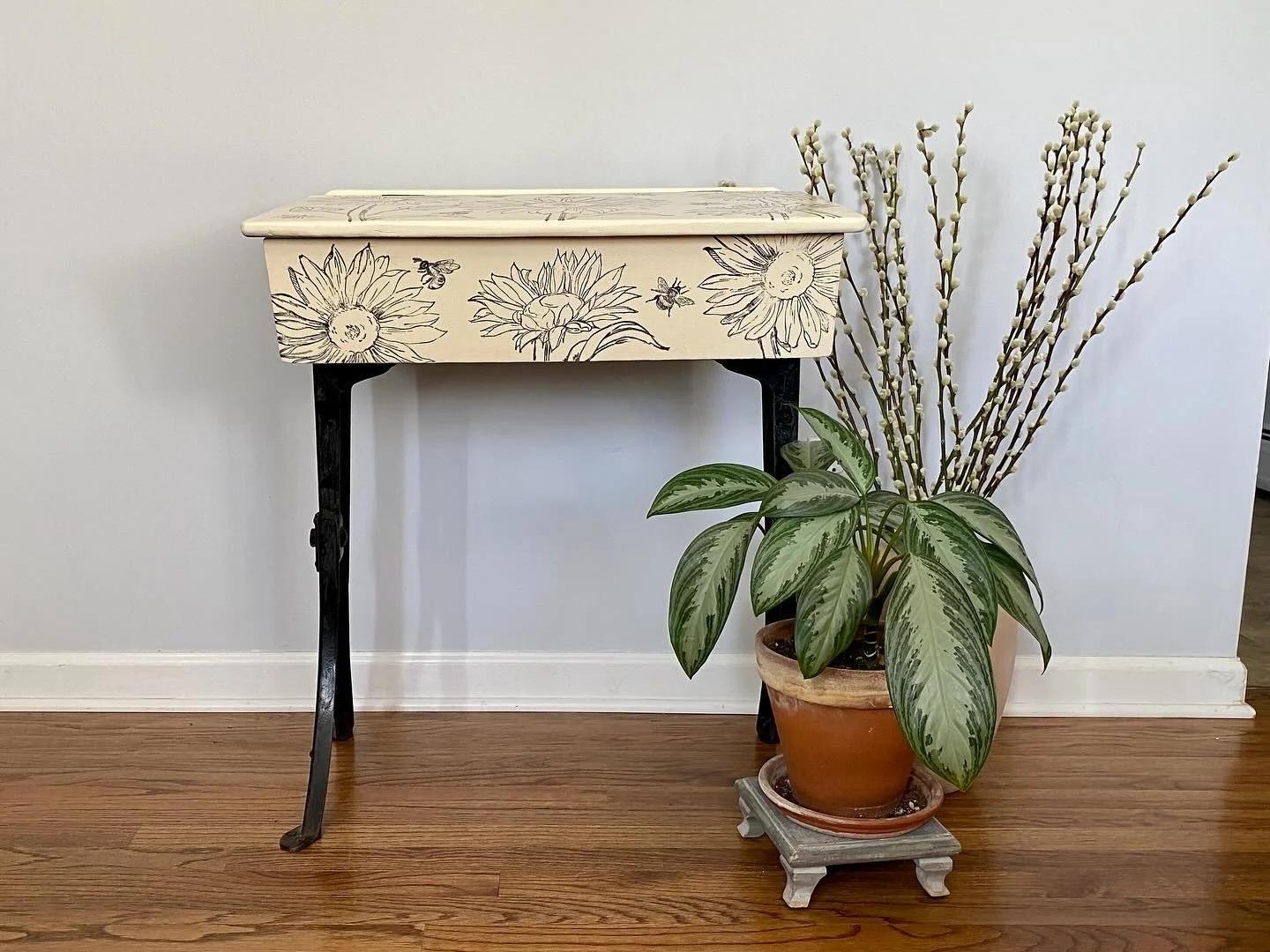 A small table with black legs and a floral design on the top and front, next to a potted plant with large green leaves and white flowers on tall stems in a terracotta pot on a small wooden stand.