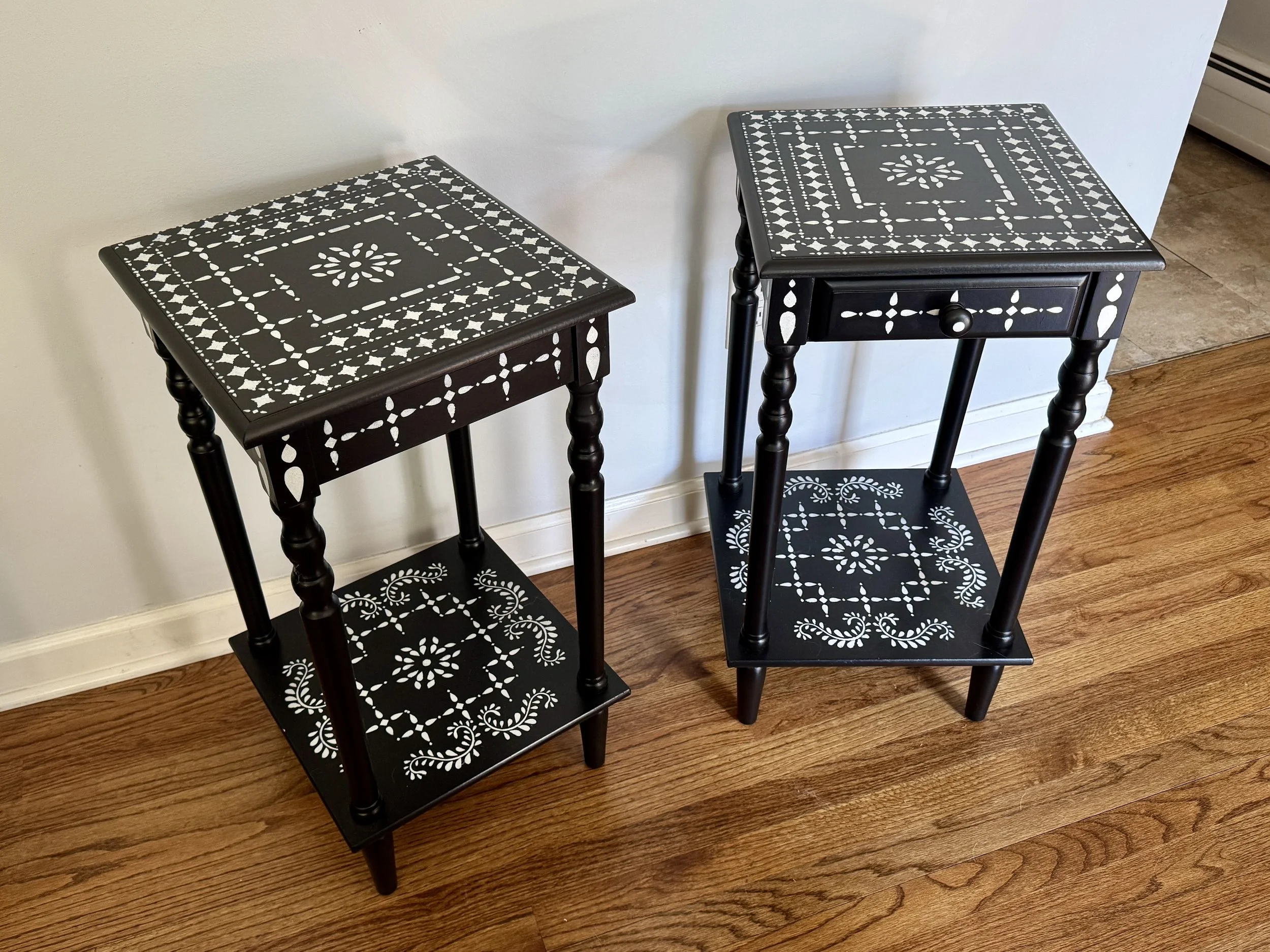 Two black wooden side tables with white decorative patterns on the tabletops and lower shelves, placed on a hardwood floor next to a white wall.