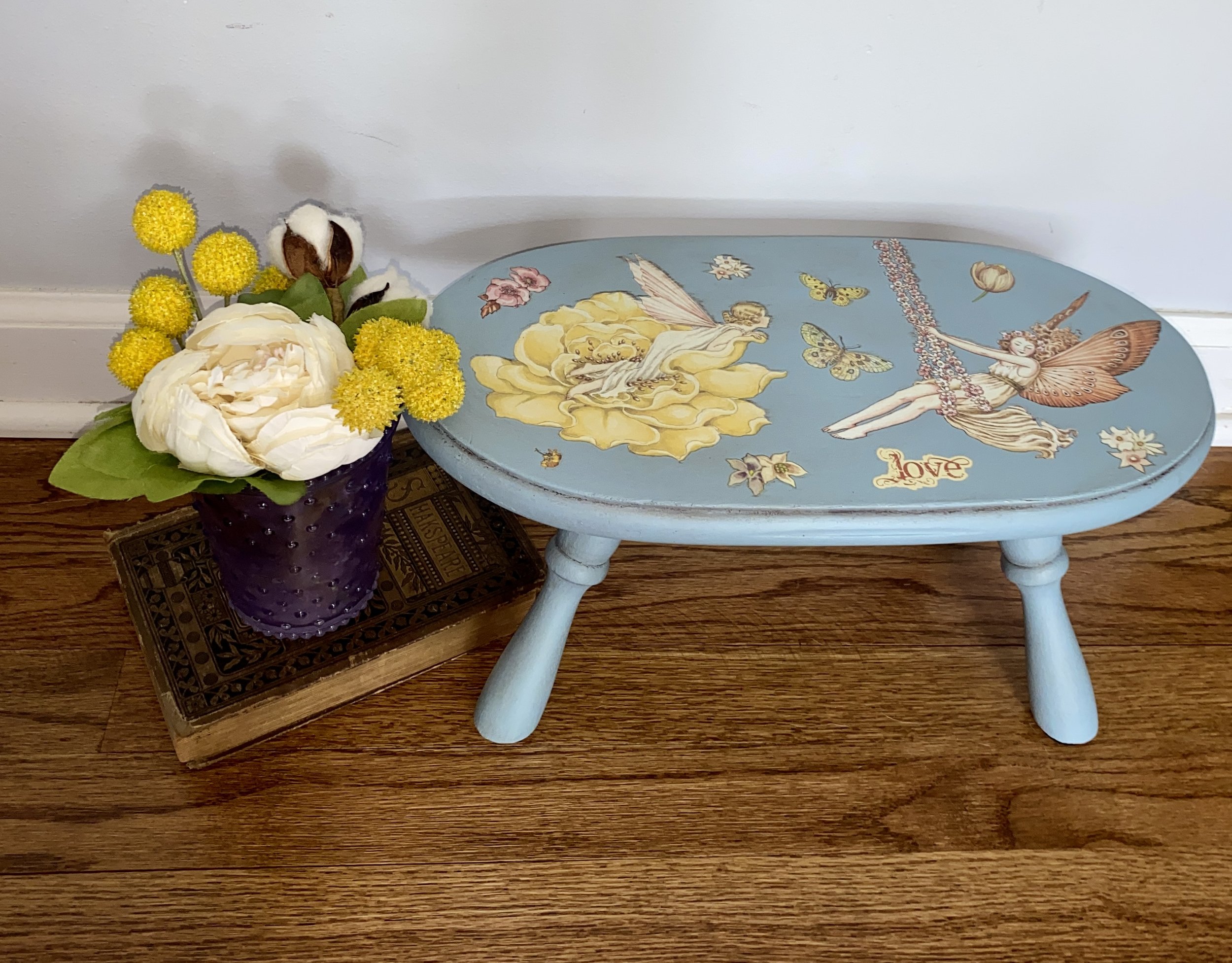 A small wooden table with a bouquet of yellow and white flowers in a purple vase, placed on top of a decorative mat. Next to the table is a painted wooden stool featuring fairy and floral illustrations, with the word "Love" written on it.