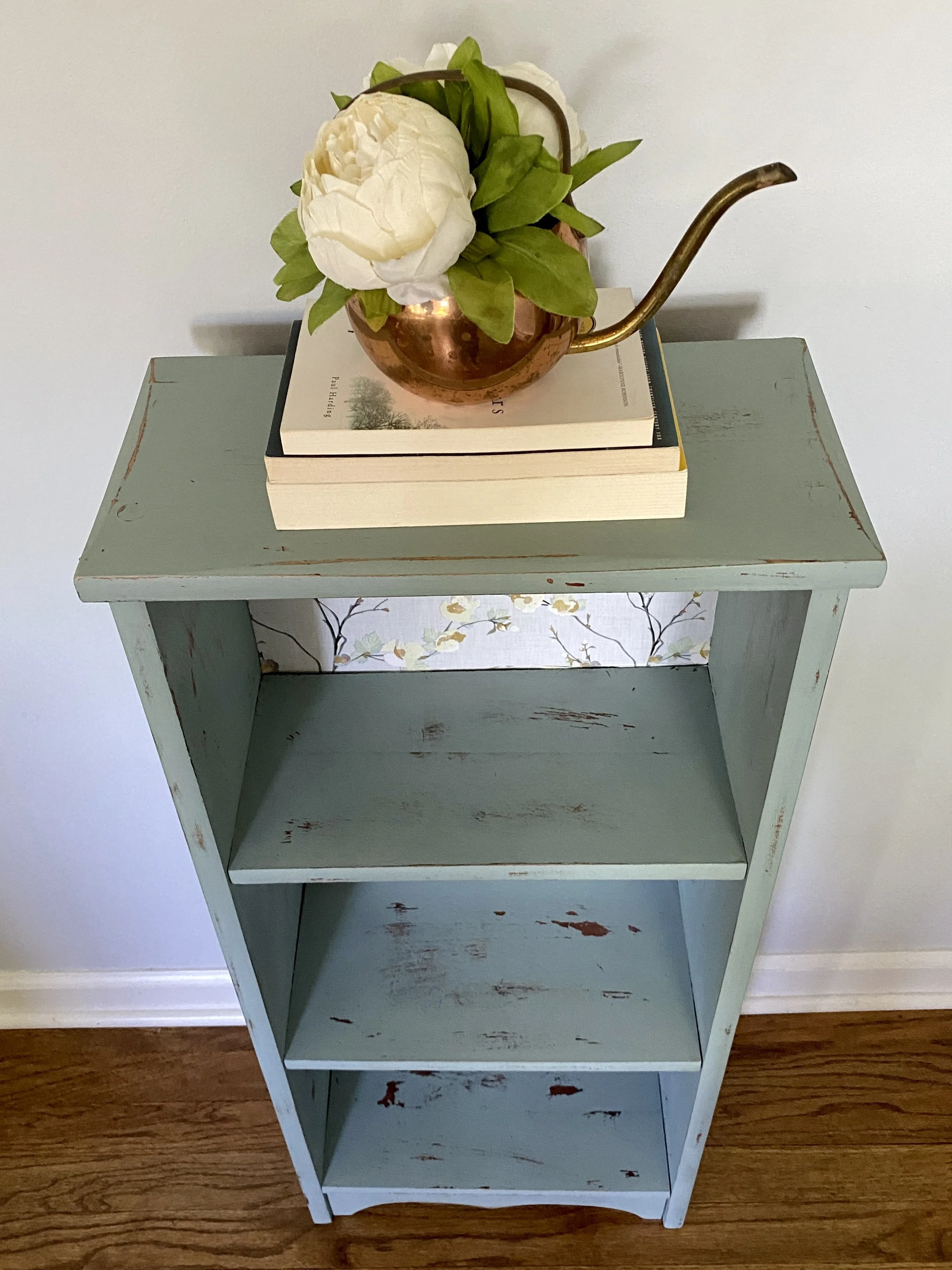 A light blue wooden bookshelf with three shelves, distressed with red paint peeks, on a hardwood floor. The top shelf holds a copper watering can with a white flower and green leaves, placed on top of two closed books.