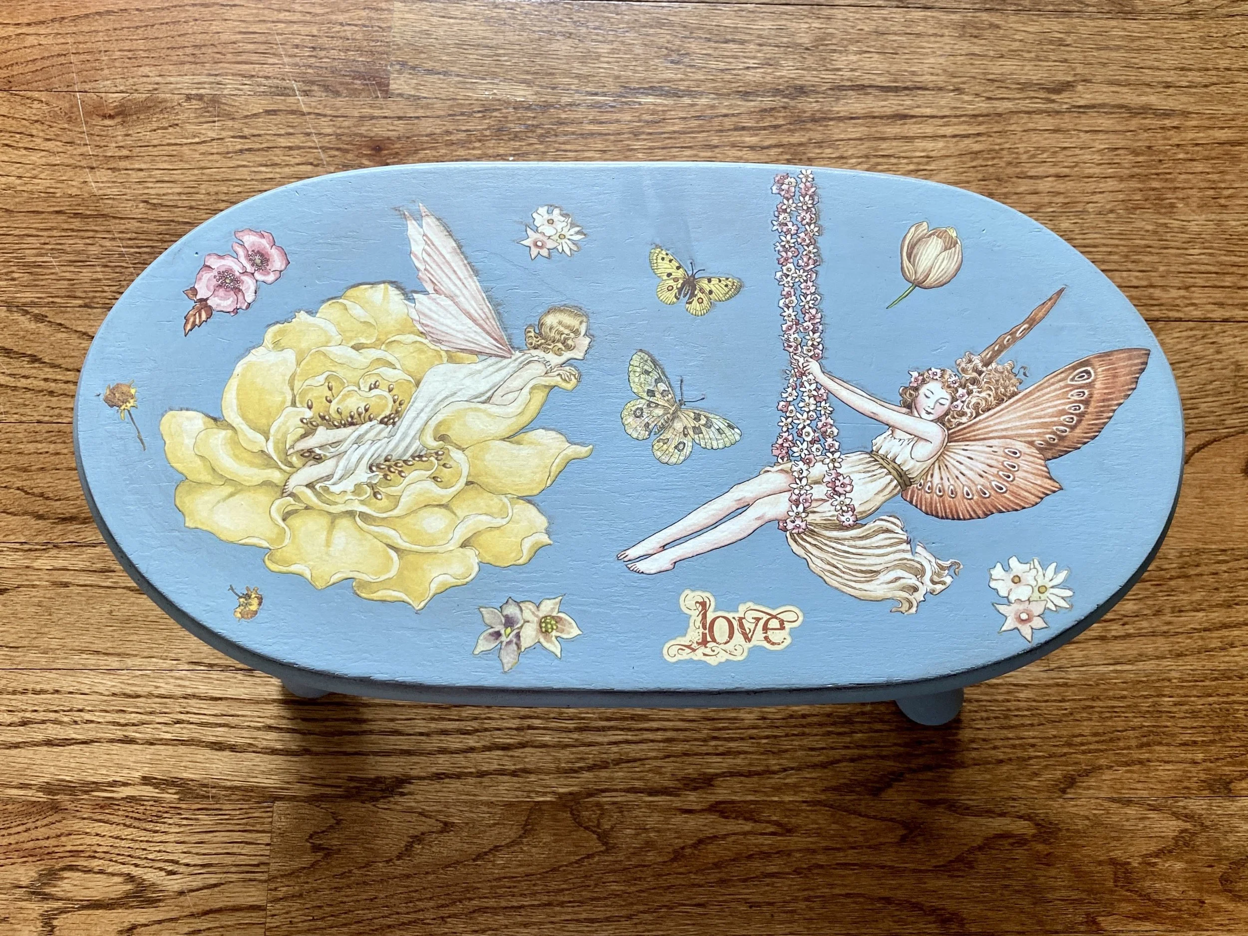Decorative oval box featuring illustrations of fairies, butterflies, flowers, and the word 'love' on a wooden surface.
