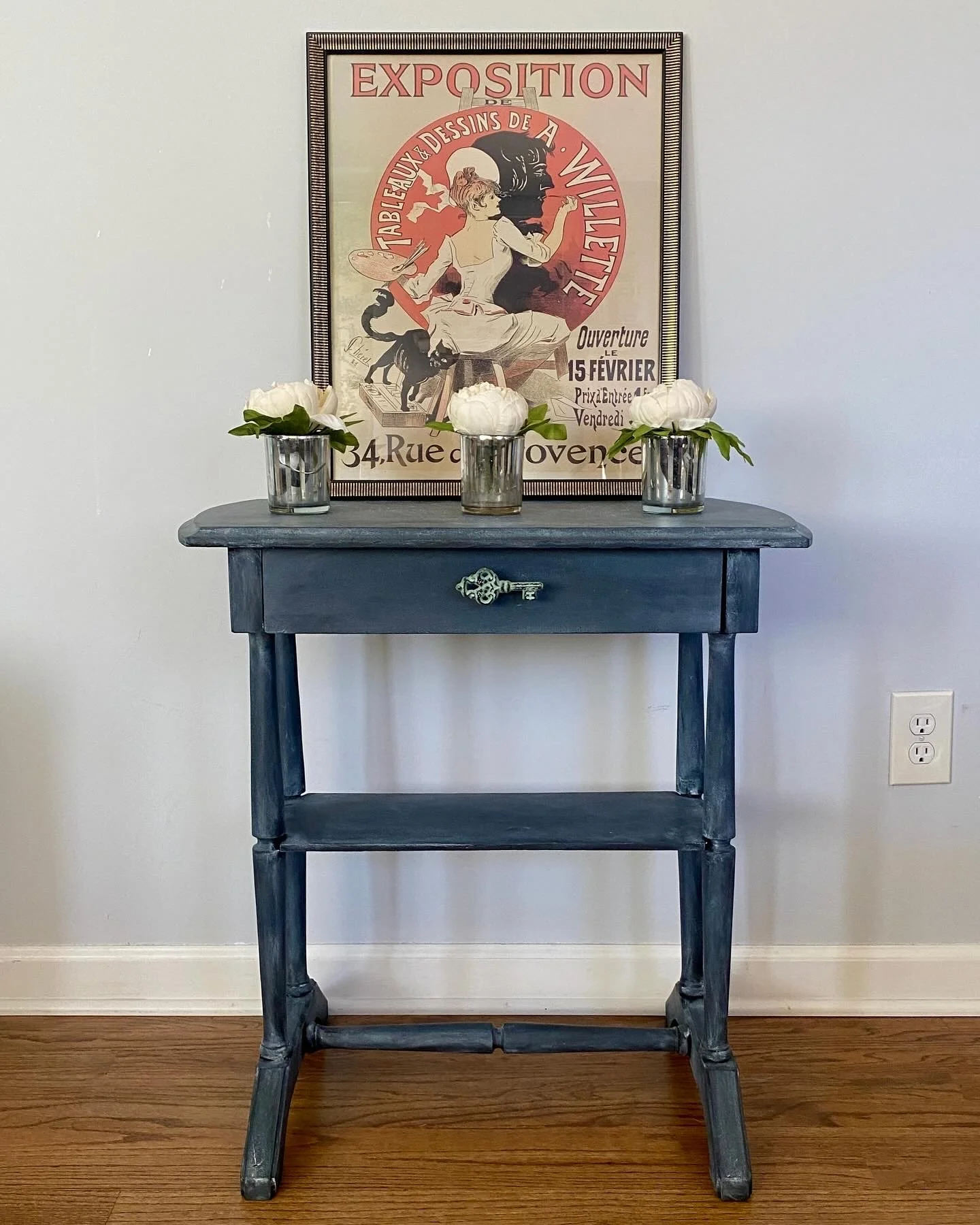 A small dark blue wooden table with a single drawer and a decorative key handle, holding three small glass vases with white flowers and green leaves, against a plain white wall with a framed vintage French poster and a white electrical outlet.
