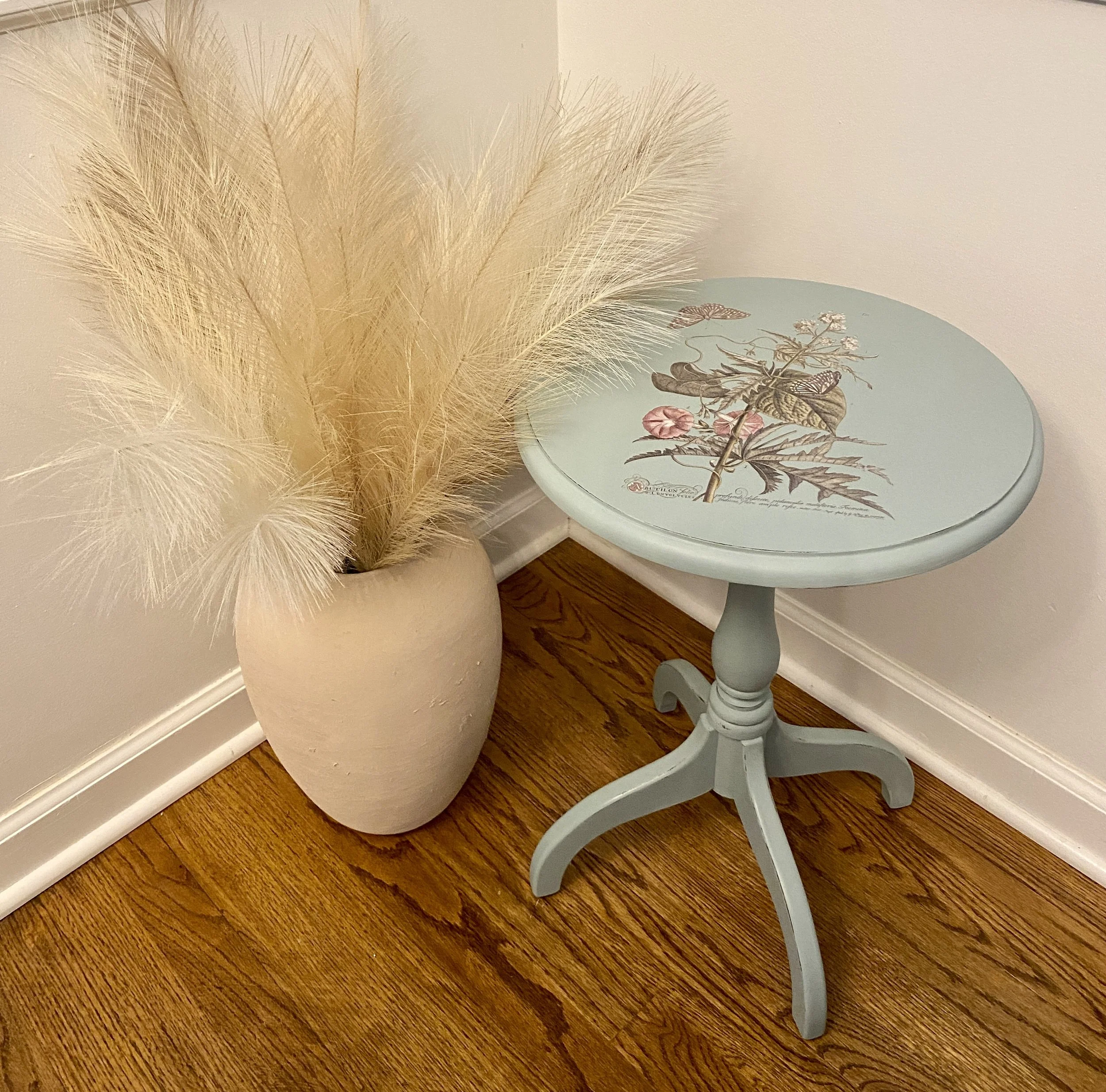A light blue round table with a botanical illustration on the top, next to a large beige vase filled with dried beige pampas grass, all placed on a wooden floor against a plain wall.