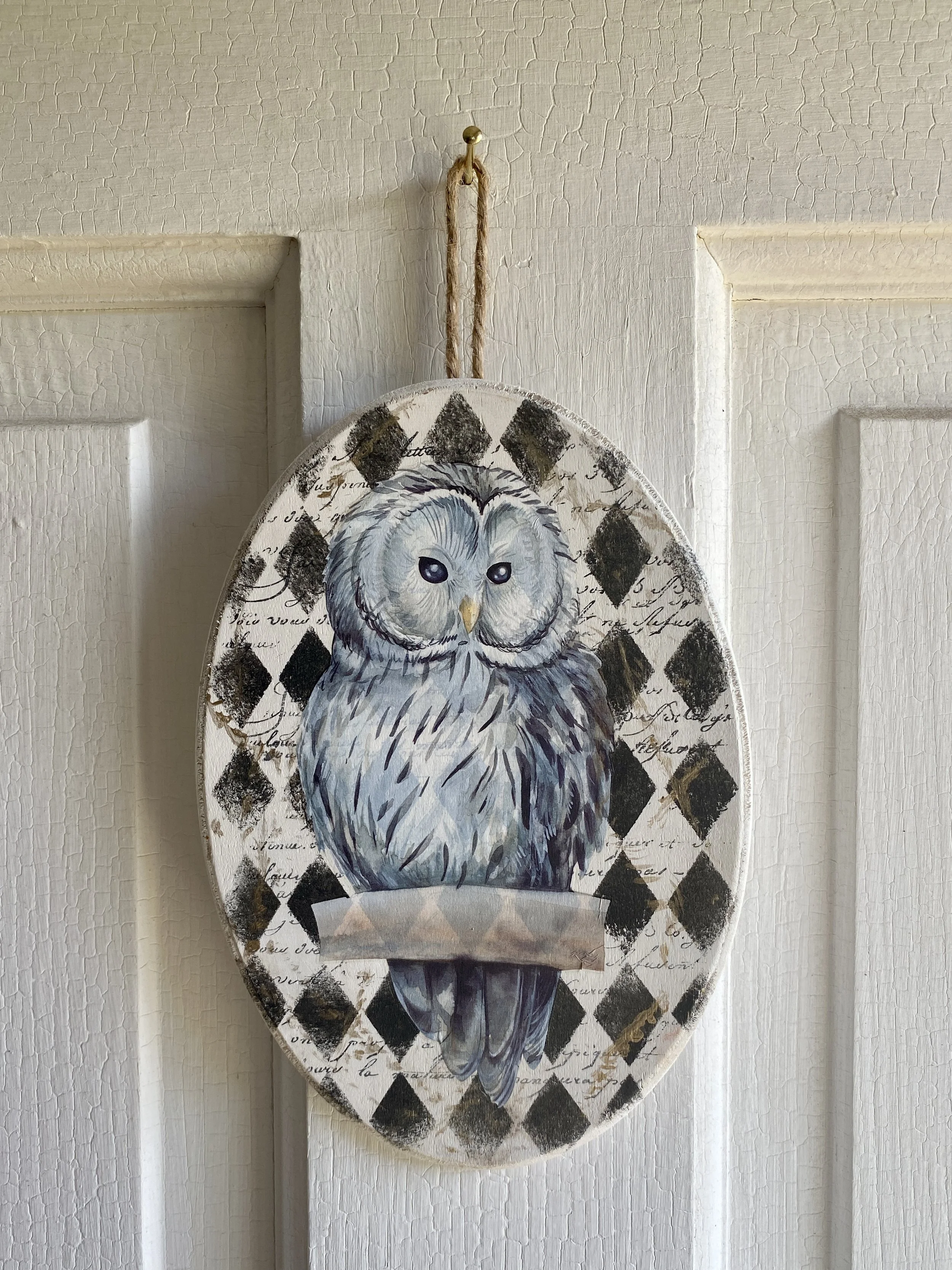 Decorative wall hanging with a painted owl on a background with handwritten script and black diamond shapes, hanging on a cream-colored wall.
