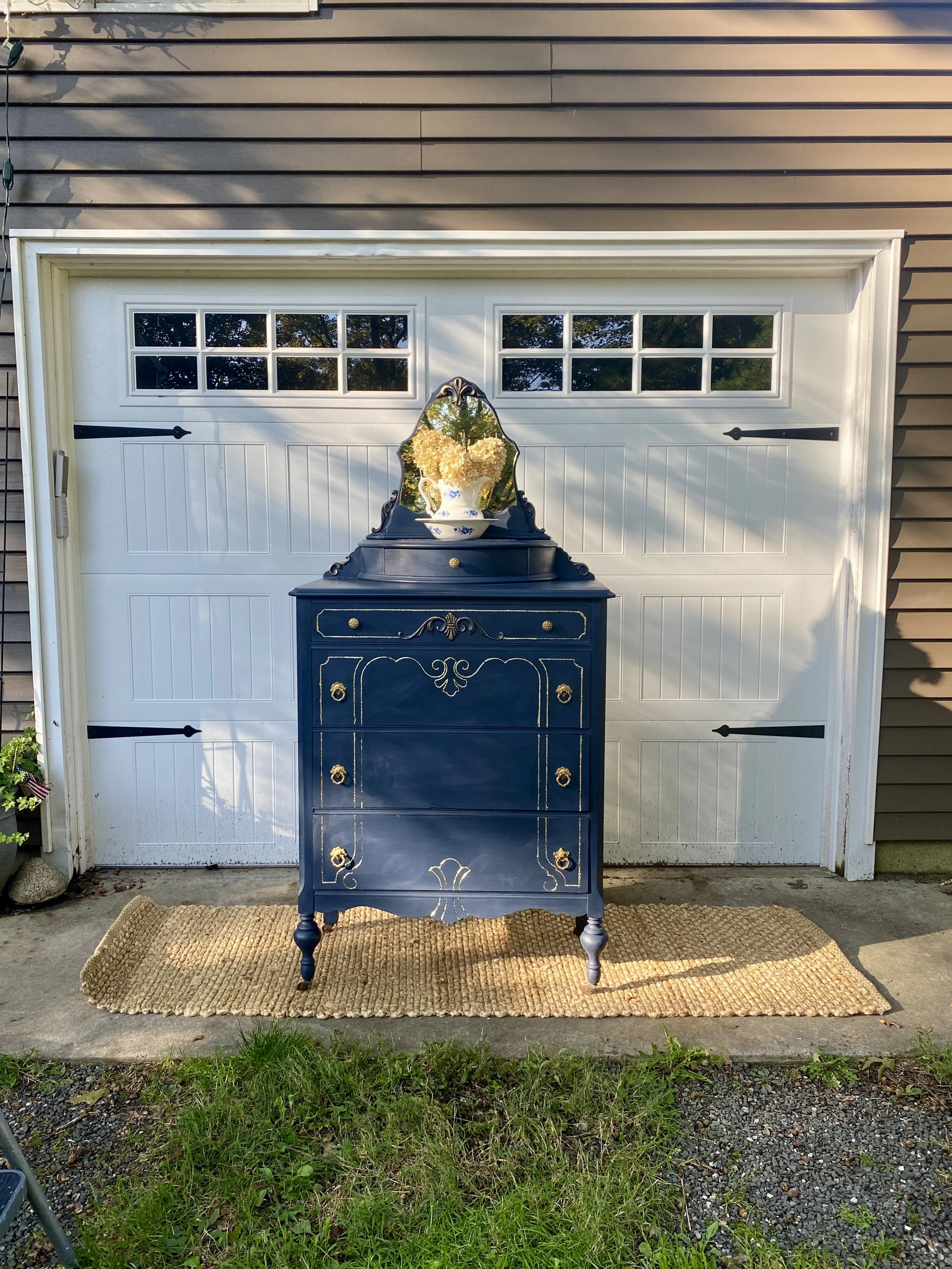 A dark blue vintage dresser with ornate gold hardware and decorative accents, placed outdoors on a woven rug in front of a brown garage door with a small mirror with a black ornate frame reflecting the trees and sky.