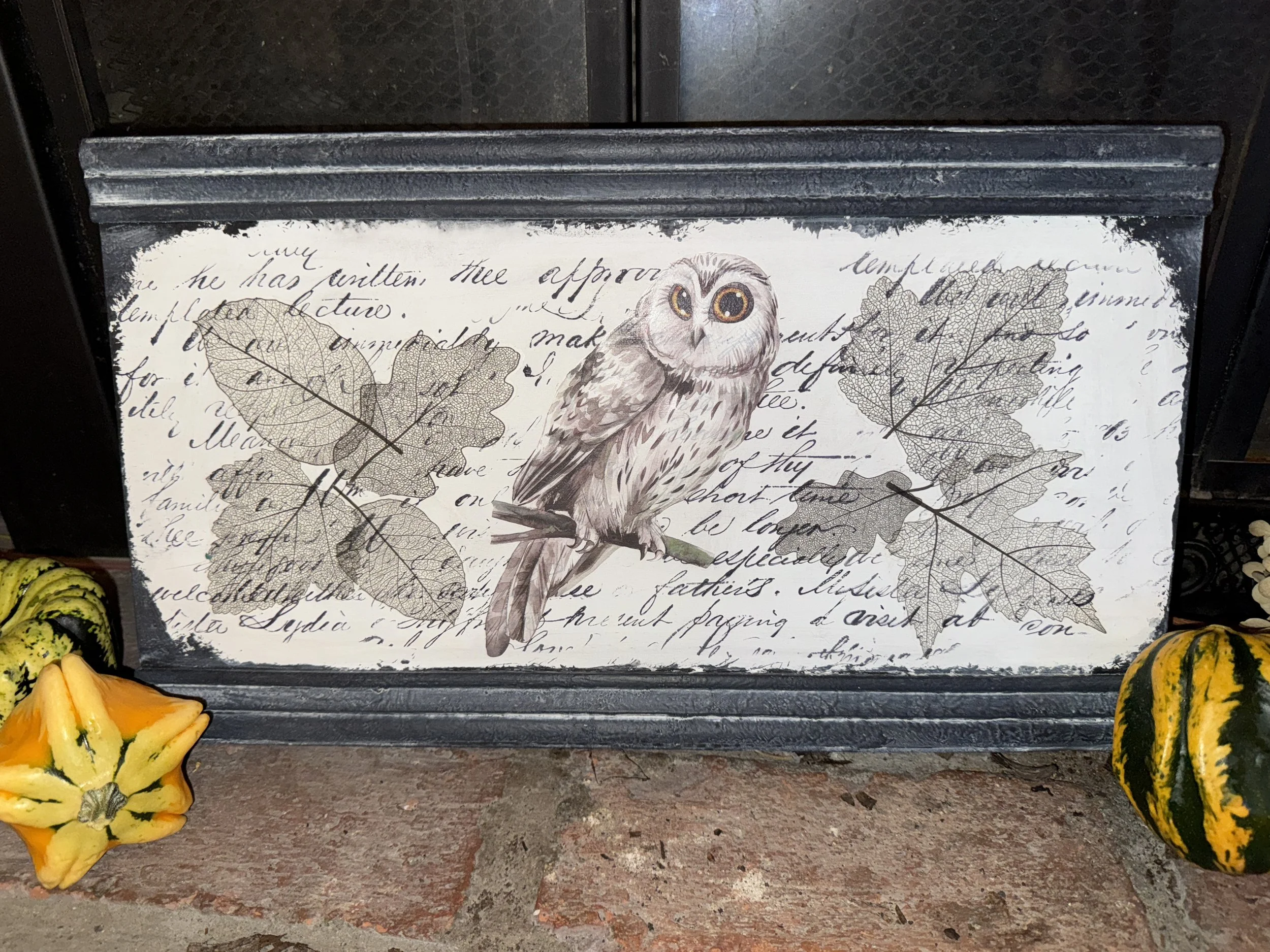 Decorative tile with an illustration of an owl perched on a branch, surrounded by autumn leaves and cursive writing in the background, placed on a shelf with gourds around it.