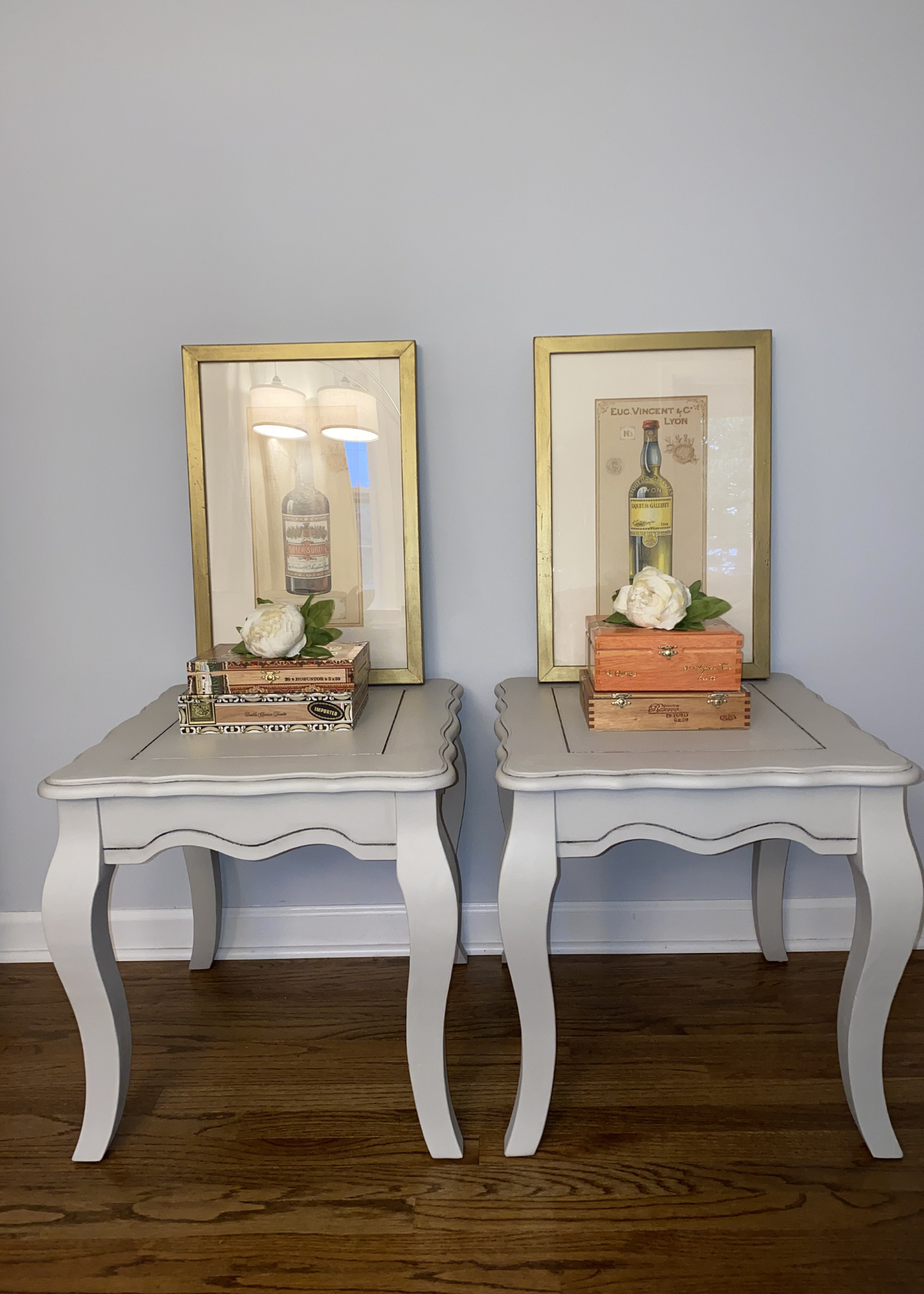 Two white vintage-style side tables with curved legs, each with a framed vintage print of a liquor bottle hanging above, decorated with white peonies and stacked decorative wooden boxes on top.