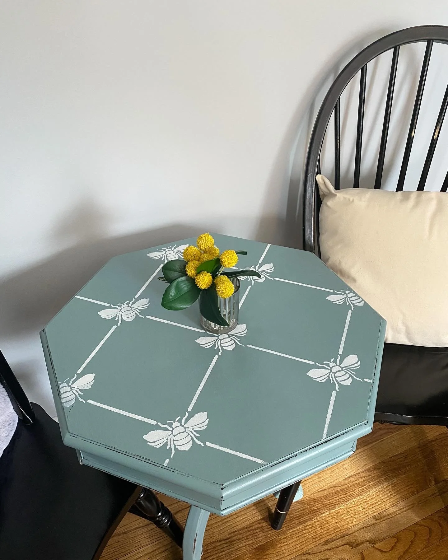 A light blue octagonal table with white bee and honeycomb pattern, topped with a small vase of yellow flowers with dark green leaves, situated next to a black chair and a beige cushion, with a white wall in the background.