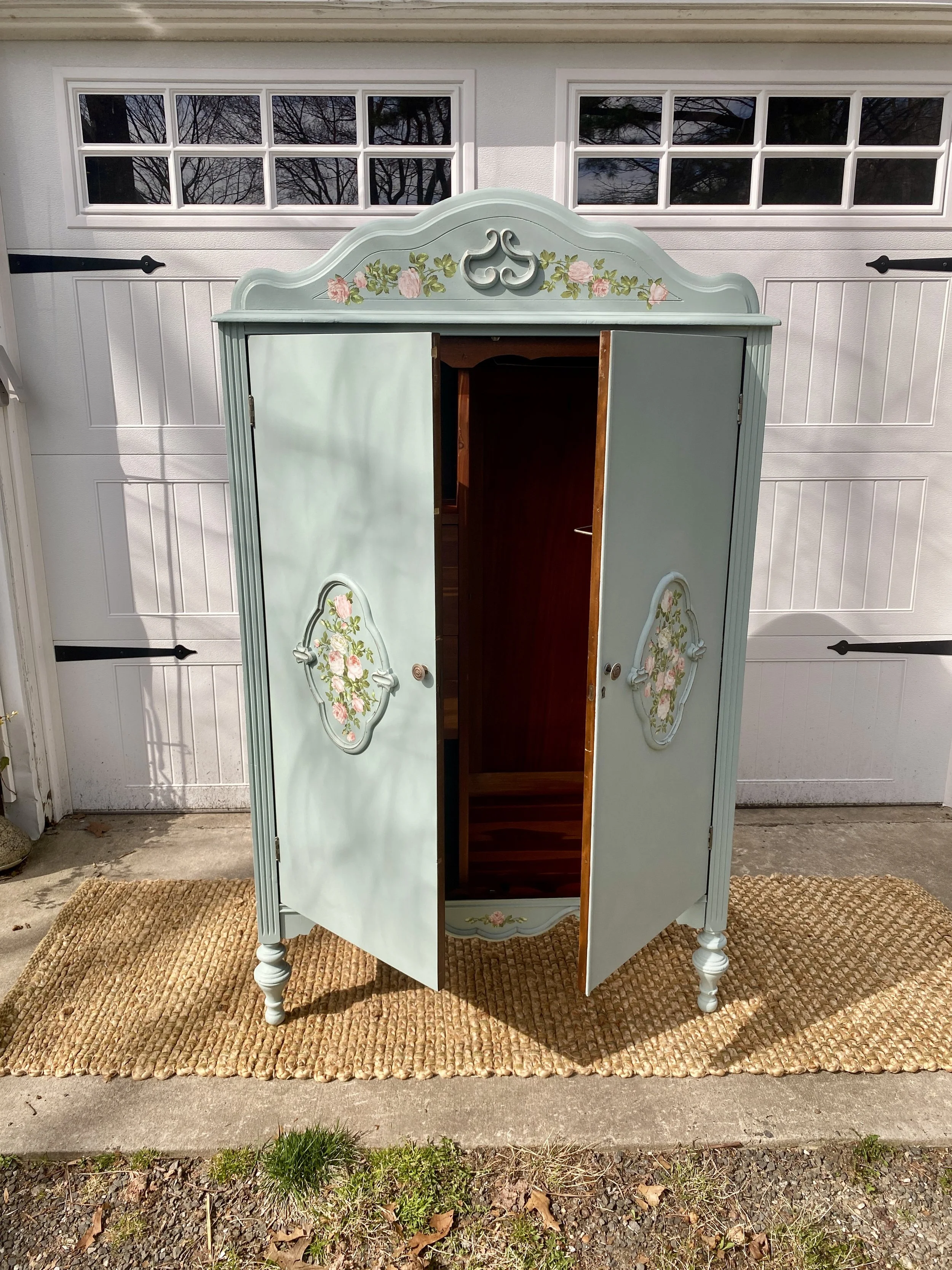 A vintage light blue painted wooden wardrobe with floral designs on the doors, standing outdoors on a brown doormat in front of a white garage door with windows.