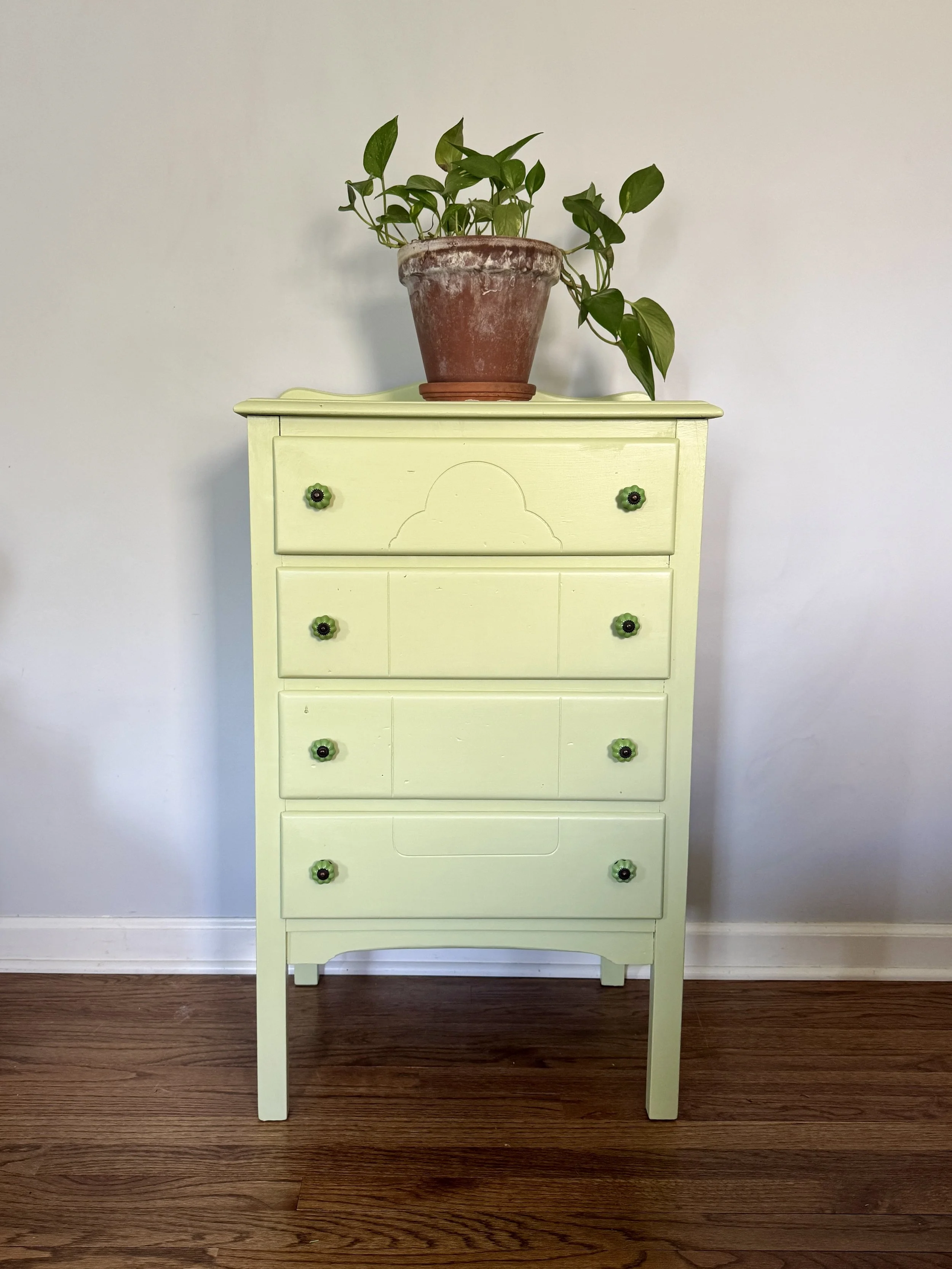 A pale yellow vintage dresser with five small drawers equipped with green and black flower-shaped knobs, standing on wooden flooring against a plain white wall. A potted plant with green leaves is placed on top of the dresser.