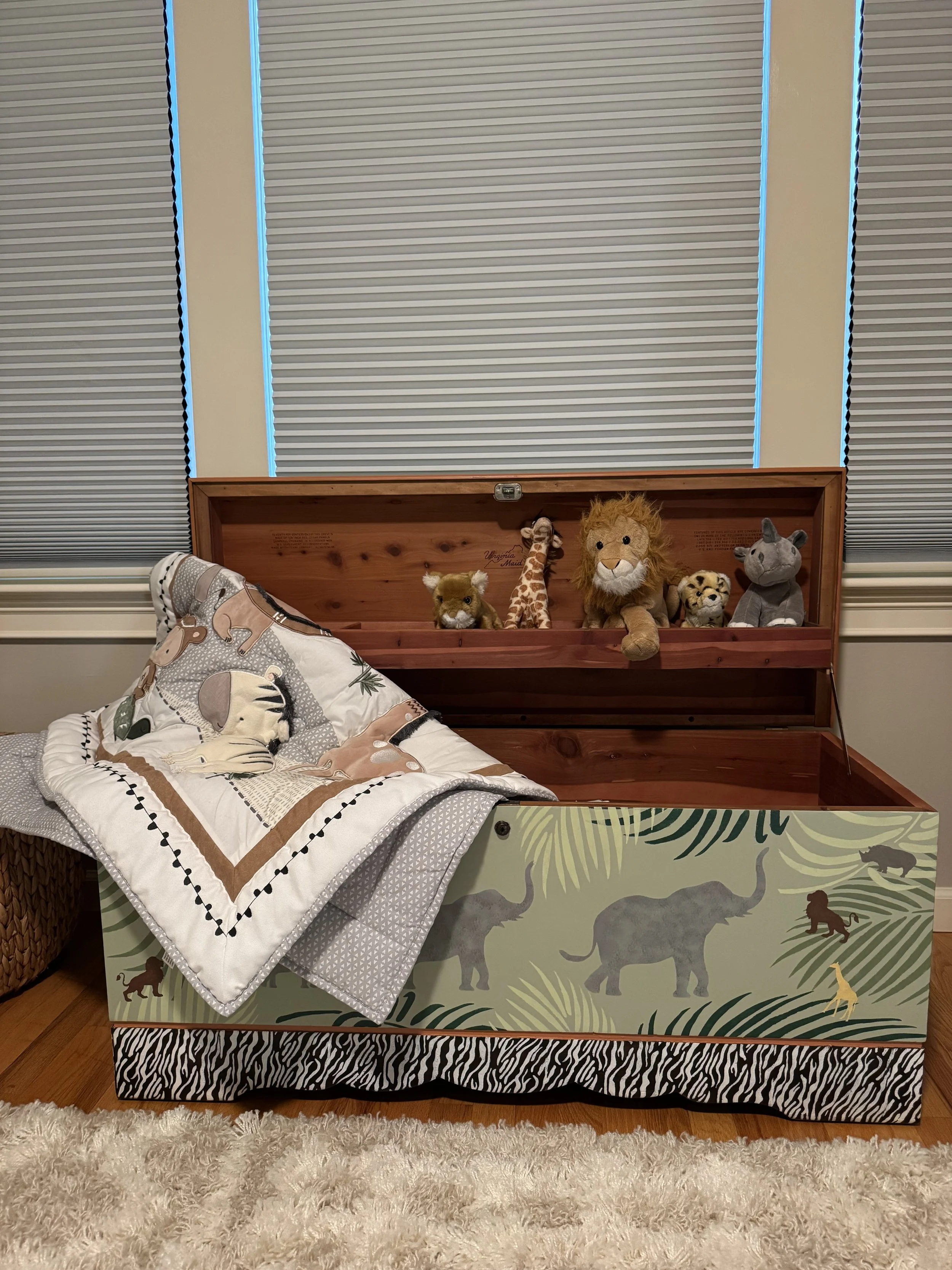 A wooden toy chest with jungle animal illustrations, stuffed animals including a lion, giraffe, tiger, and rhino, with a quilted blanket on bed, and closed blinds in the background.