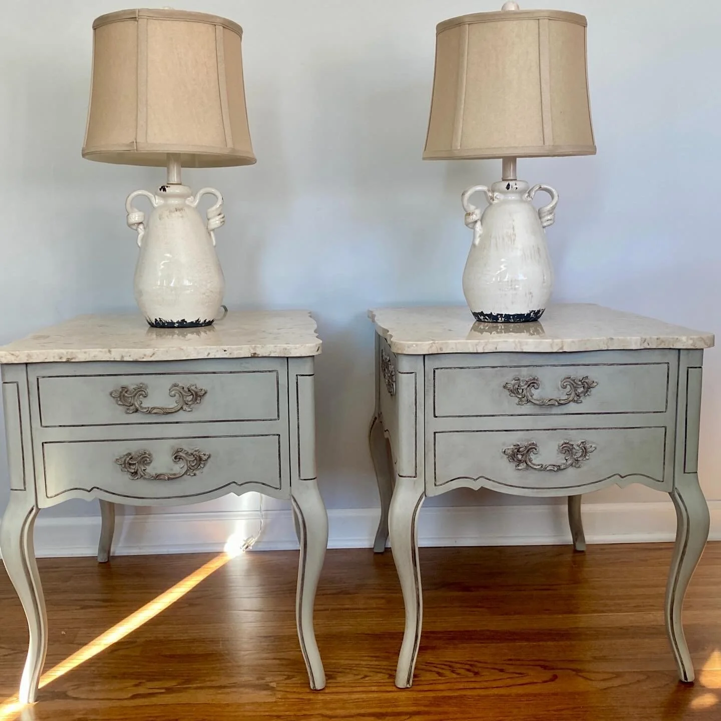 Pair of vintage-style bedside tables with white distressed finish, ornate handles, marble tops, each topped with a beige lampshade and decorative ceramic vases.