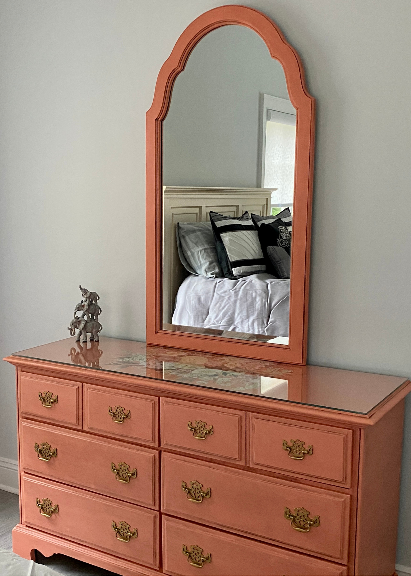 A coral-colored vintage dresser with a marble top and ornate brass drawer pulls, topped with a matching coral-framed mirror. The mirror reflects a bed with pillows and a white headboard, and a window with light-colored curtains in a bedroom.