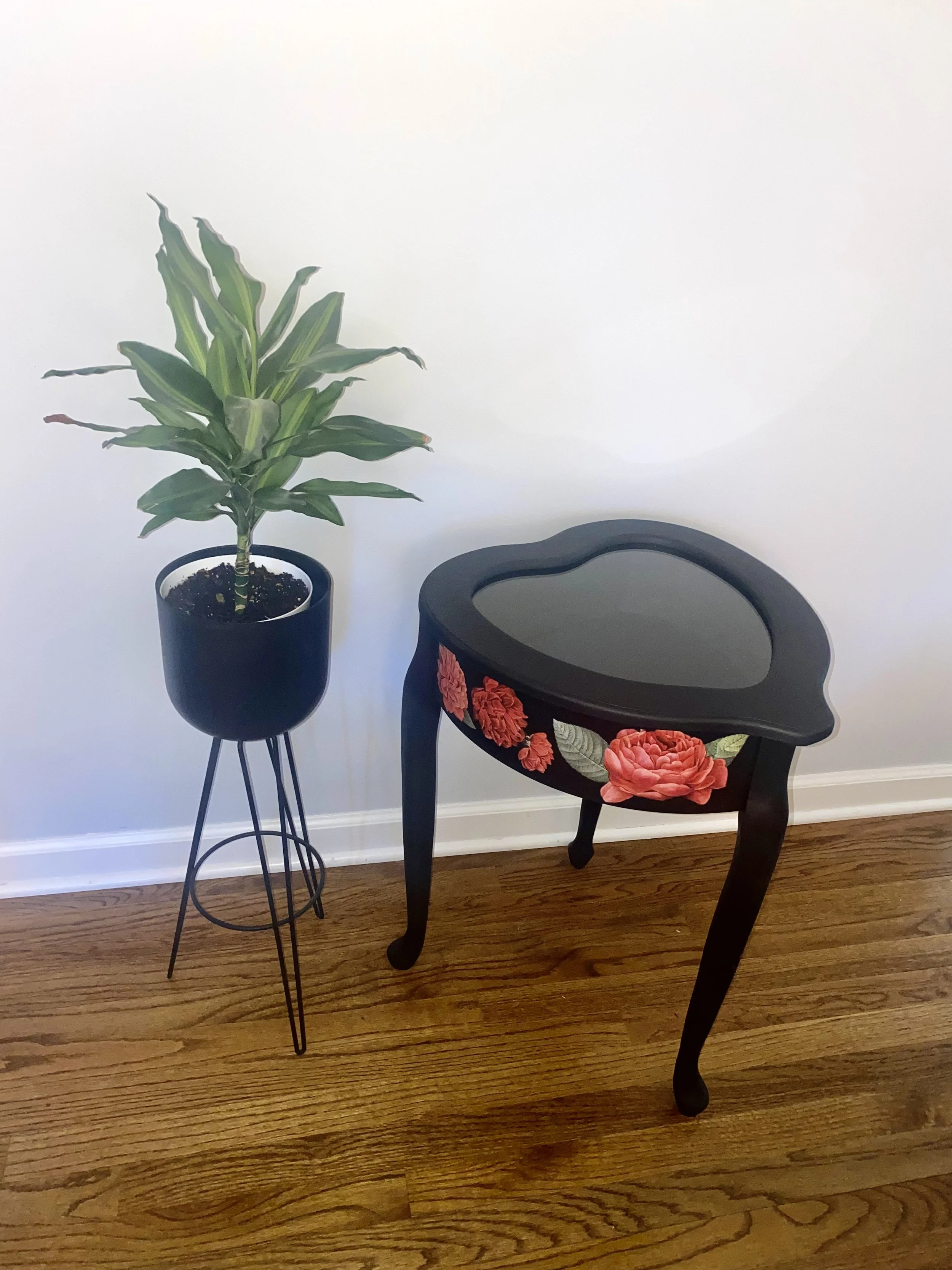 A black chair with floral carvings on its backrest, a large potted green plant in a black pot on a metal stand, and a wooden floor against a plain white wall.