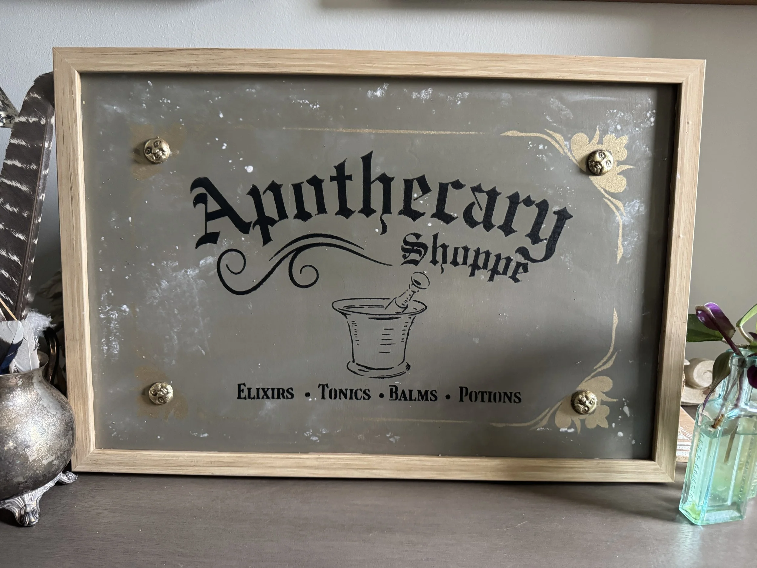 A decorative sign with a wooden frame and metallic background, featuring the word 'Apothecary' in black text, along with illustrations and words related to herbal remedies such as 'Elixirs,' 'Tonics,' 'Balms,' and 'Potions'.