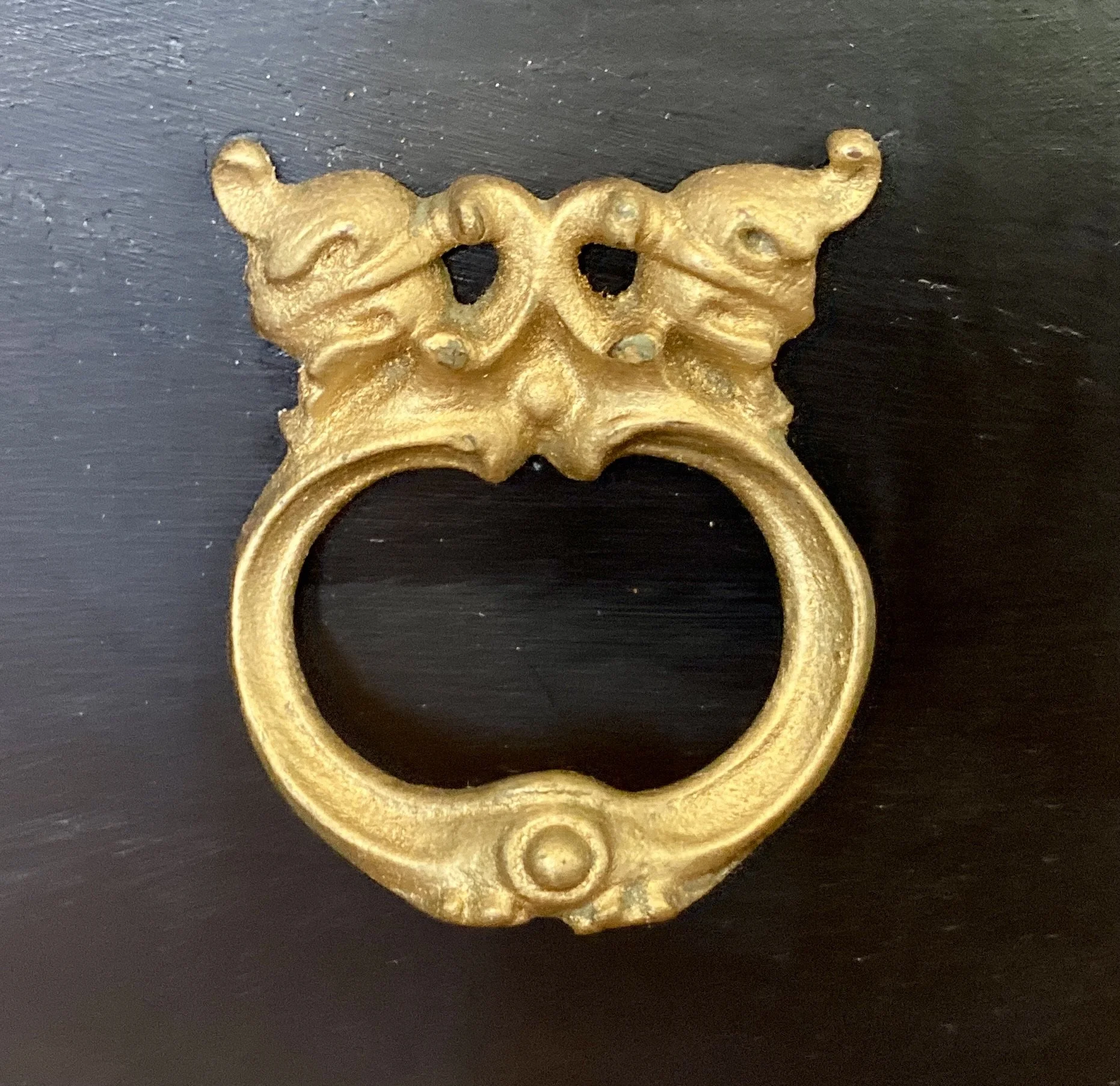 A gold decorative door knocker with intricate design, featuring a face with curled motifs and a circular ring beneath, set against a dark background.