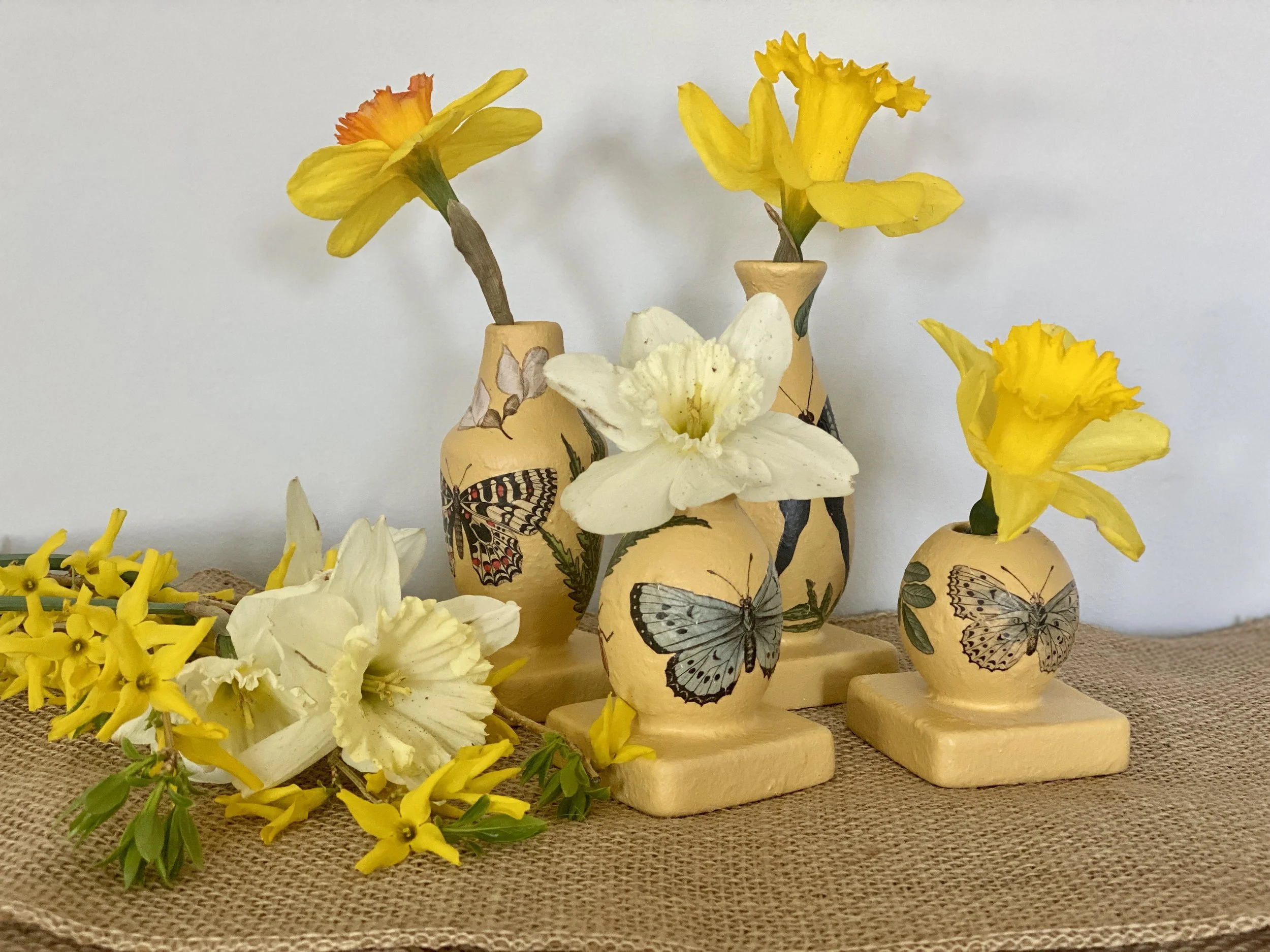 Multiple small decorative vases with painted butterfly designs containing yellow and white flowers, arranged on a woven mat with yellow flowers scattered around.