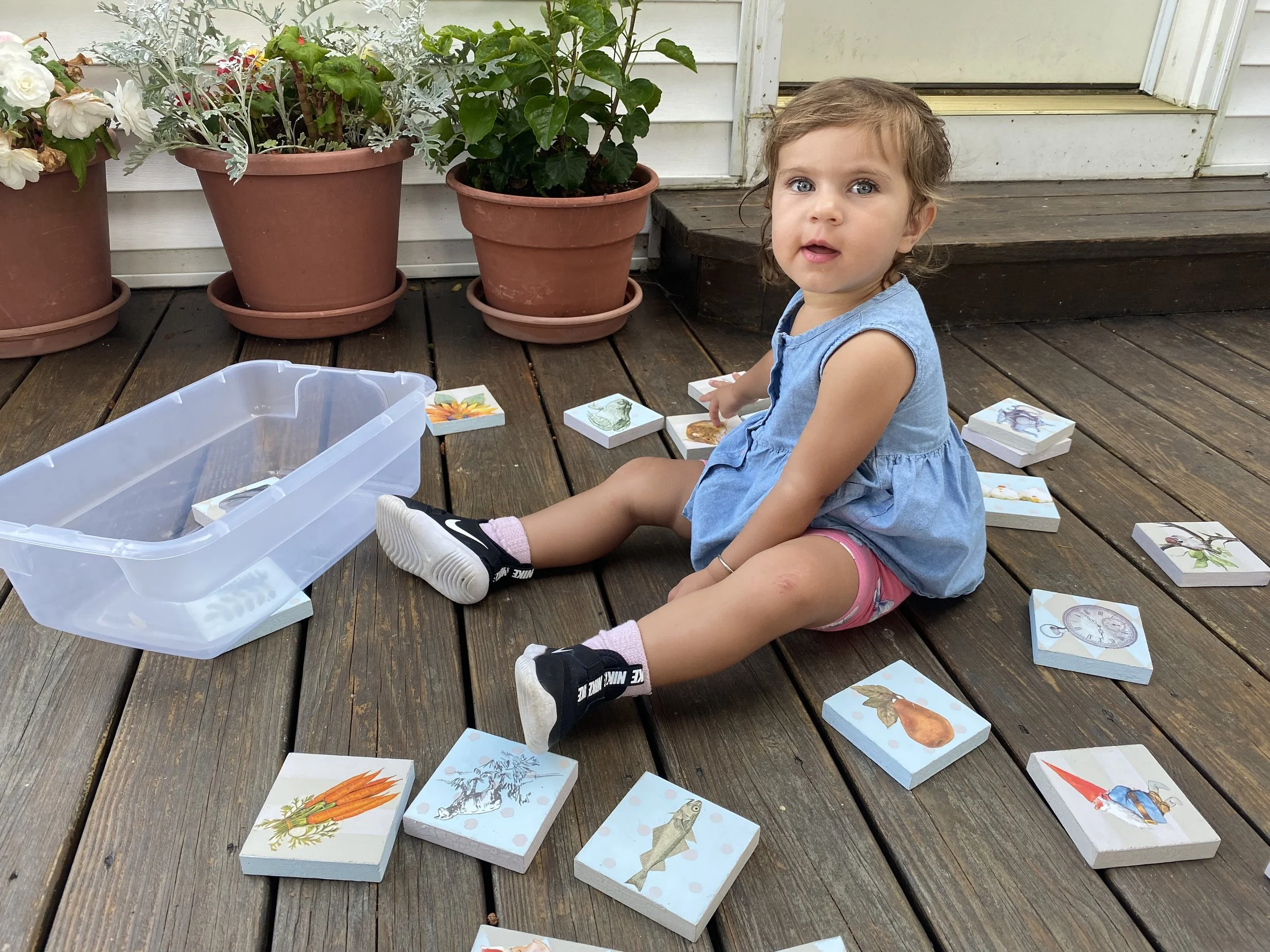 A young girl with blue eyes and brown, slightly curly hair, sitting on a wooden deck outdoors. She is wearing a blue sleeveless dress and pink shorts, with black sneakers and pink socks. Around her are various illustrated cards spread on the deck, so