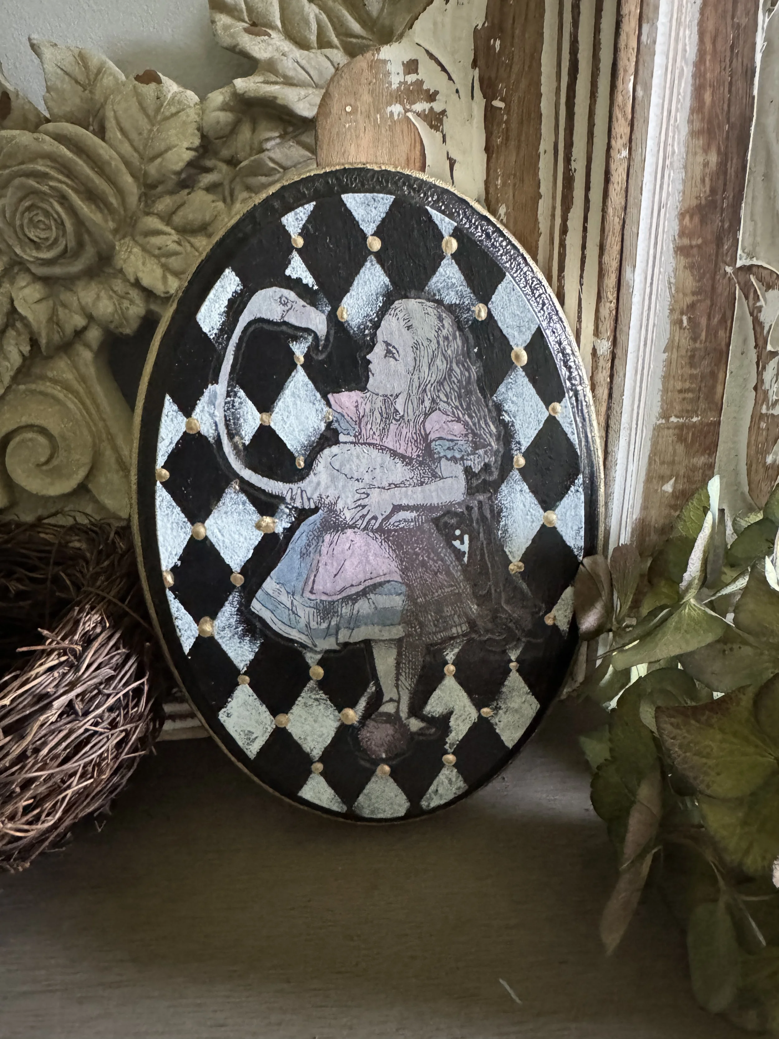 Decorative oval mirror with a black and white checkered pattern, featuring a depiction of a woman with long hair, surrounded by dried leaves and a woven basket on a wooden surface.