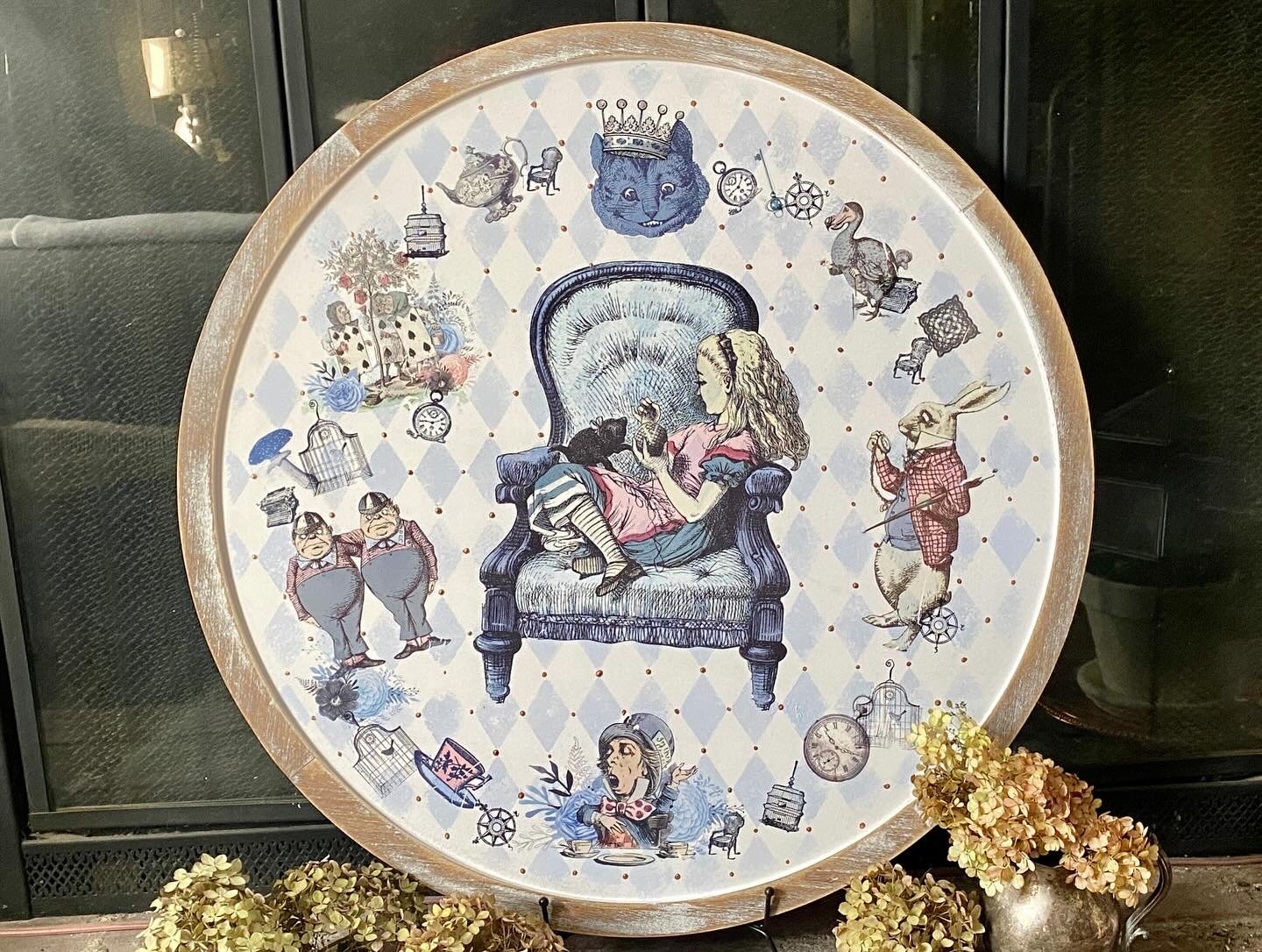 Decorative round tray with a central illustration of a girl sitting on a vintage armchair, surrounded by various whimsical characters and objects, including animals, clocks, and birdcages, all set against a light diamond-patterned background.
