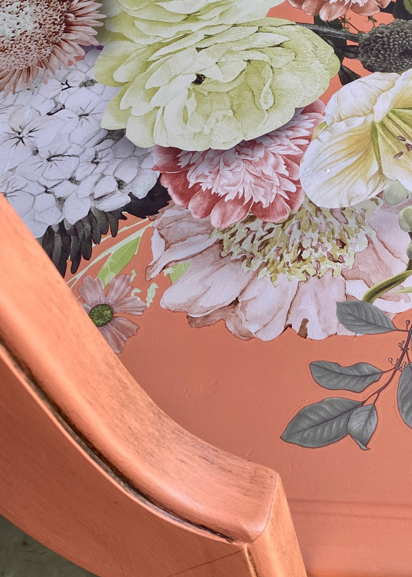 Close-up of a floral wallpaper design with large, detailed flowers in soft pastel colors, including pink, white, and yellow, and green and dark leaves, bordered by a peach-colored wooden surface.