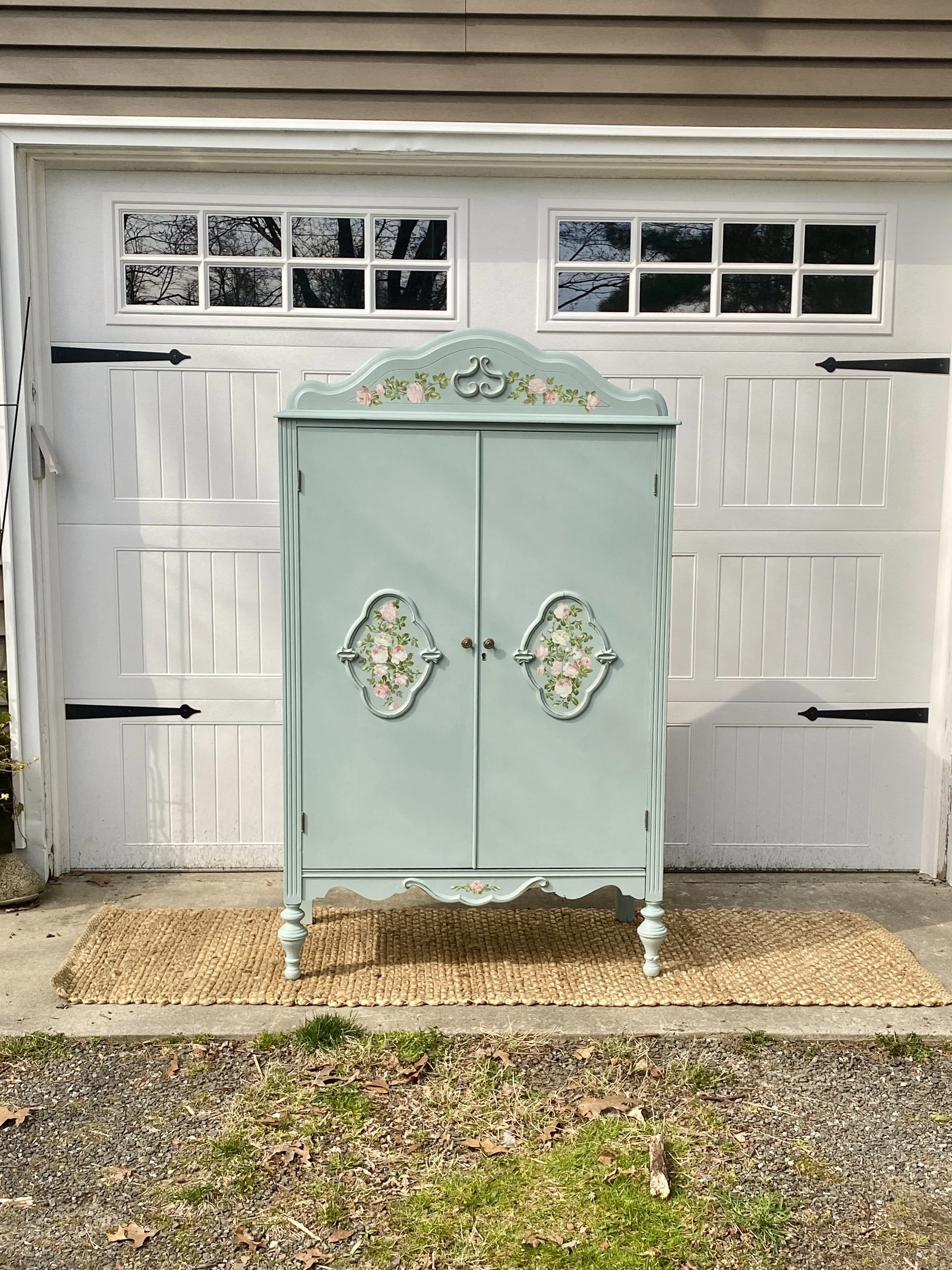 A light green vintage style cabinet with floral decorations in front of a white garage door, placed on a woven rug on a concrete porch with grass and fallen leaves.