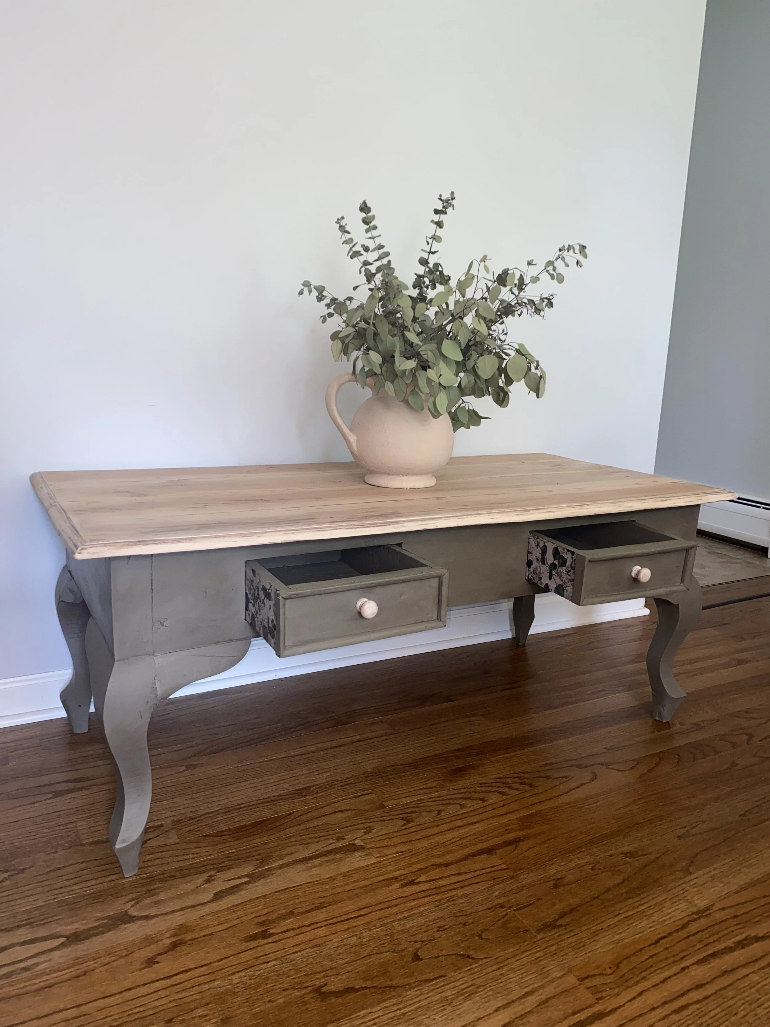 A vintage-style wooden table with carved legs and two open drawers, placed on a hardwood floor. A large beige vase with green leafy branches decorates the table.