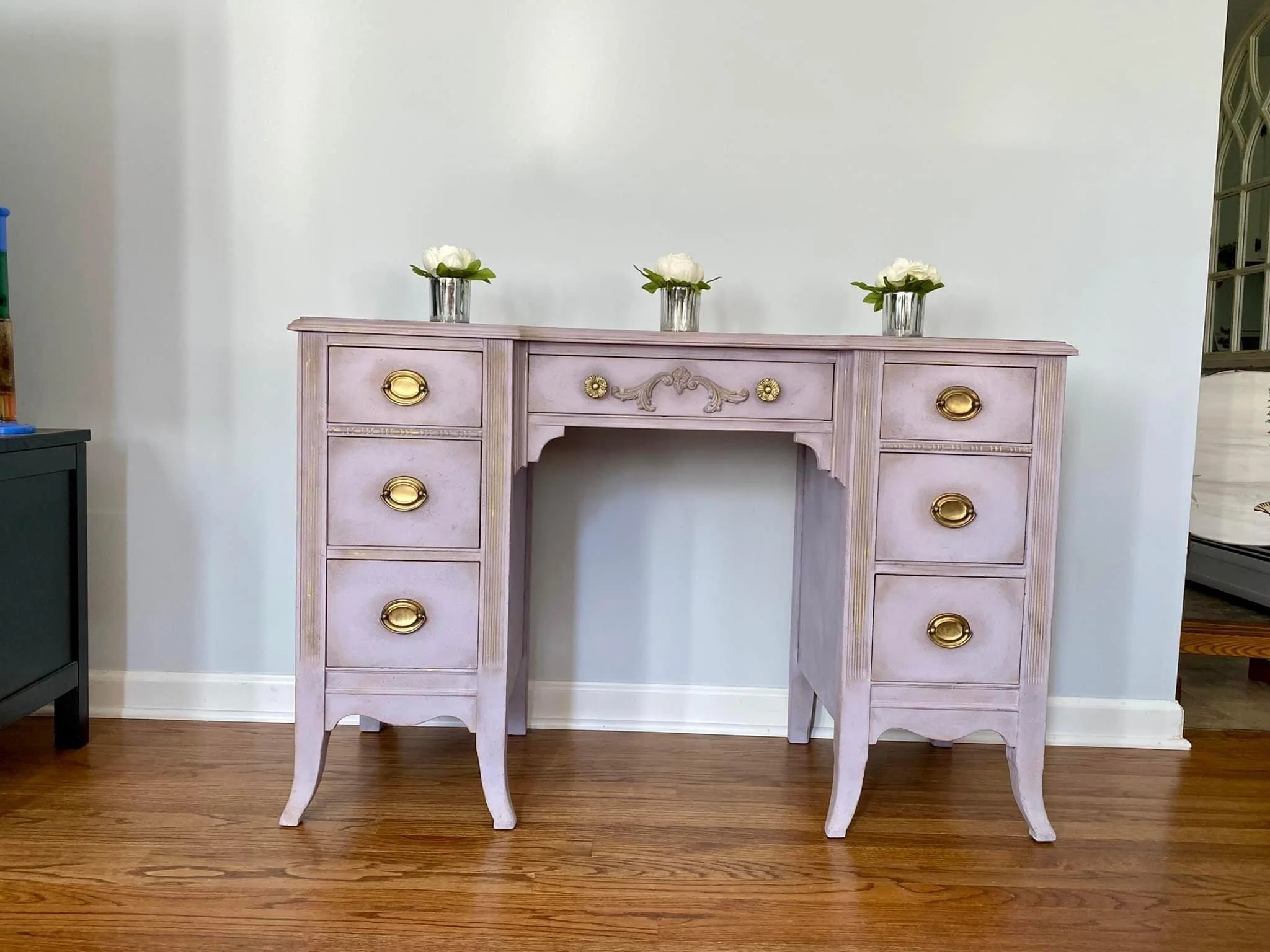 A vintage pink wooden vanity with gold drawer pulls and ornate detailing, three white flowers in glass vases on top, against a plain white wall. Part of a dark piece of furniture is visible on the left, with a mirror and part of a doorway to the righ
