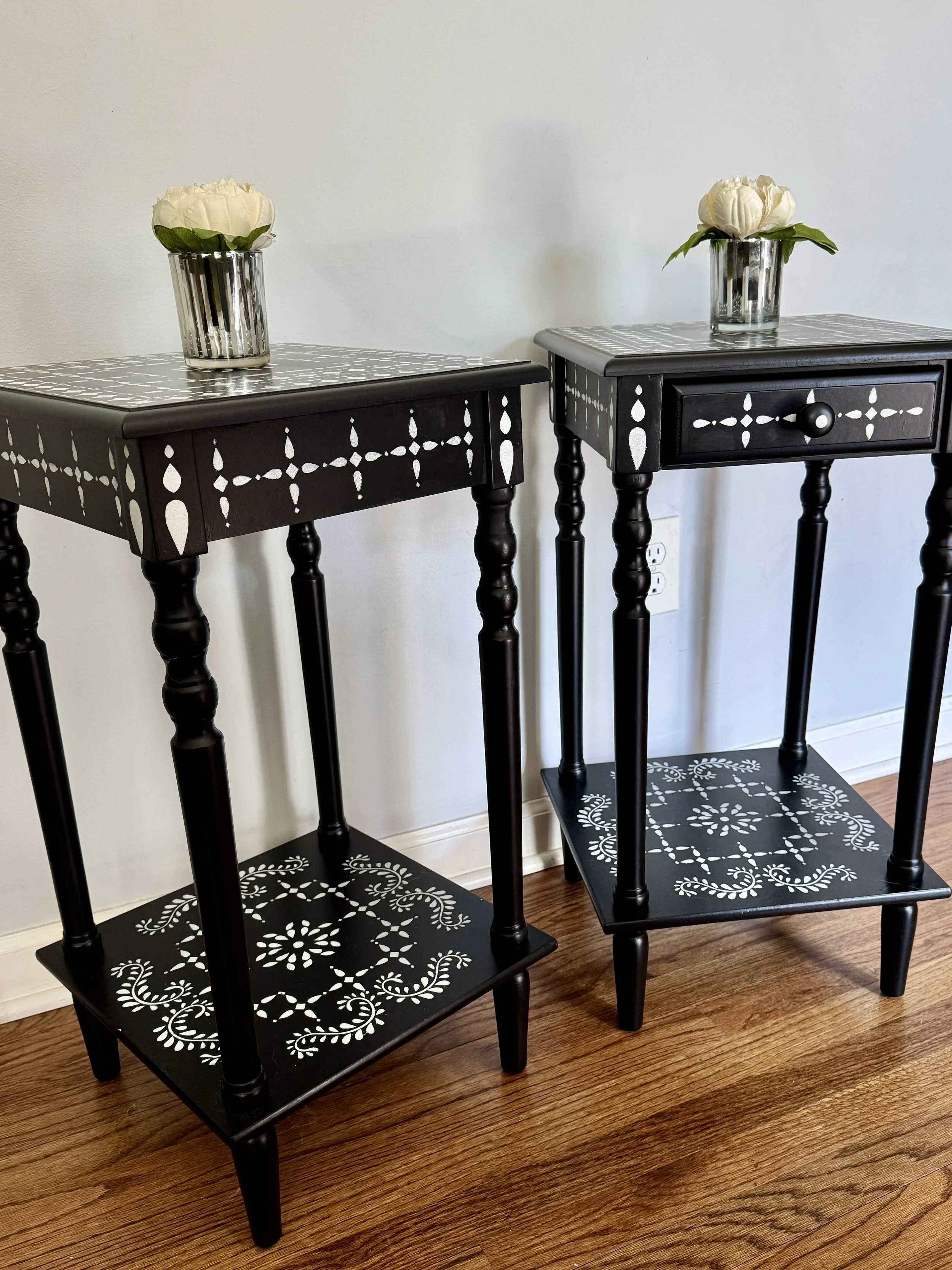Two black wooden side tables with white painted decorative patterns, each with a glass vase containing white peonies and green leaves on top, placed on hardwood flooring against a plain white wall.