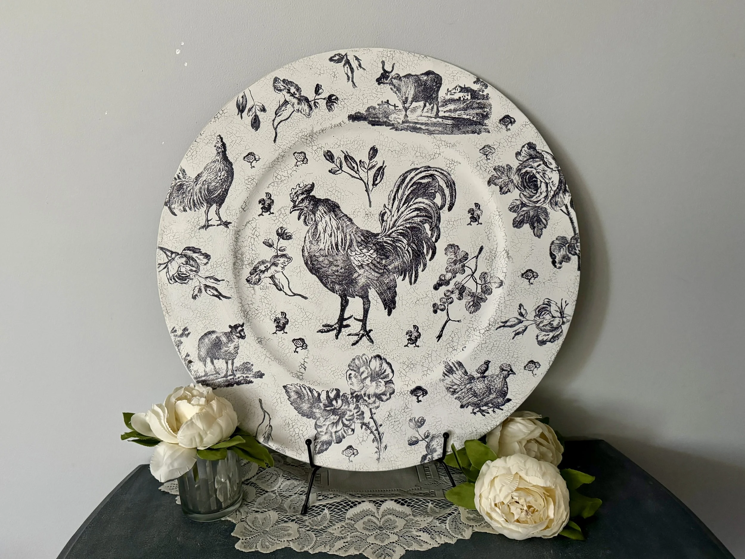 A decorative plate with a vintage black-and-white farm scene featuring chickens, a rooster, and a cow, set on a stand surrounded by white flowers and placed on a table with a lace tablecloth against a plain wall.