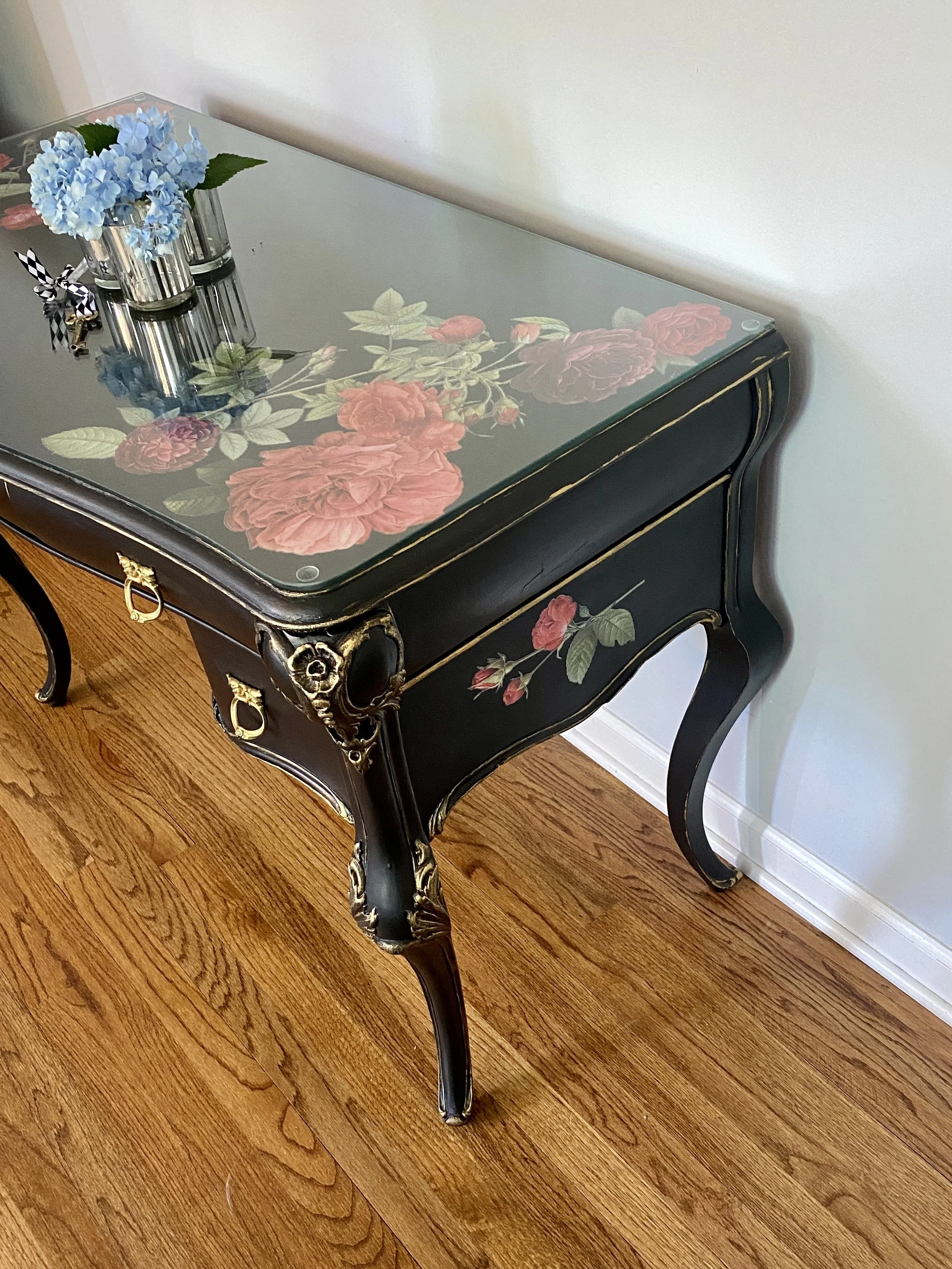 Black vintage side table with floral and leaf pattern on top, topped with three small glass vases with blue hydrangeas and green leaves, on a wooden floor with baseboard.