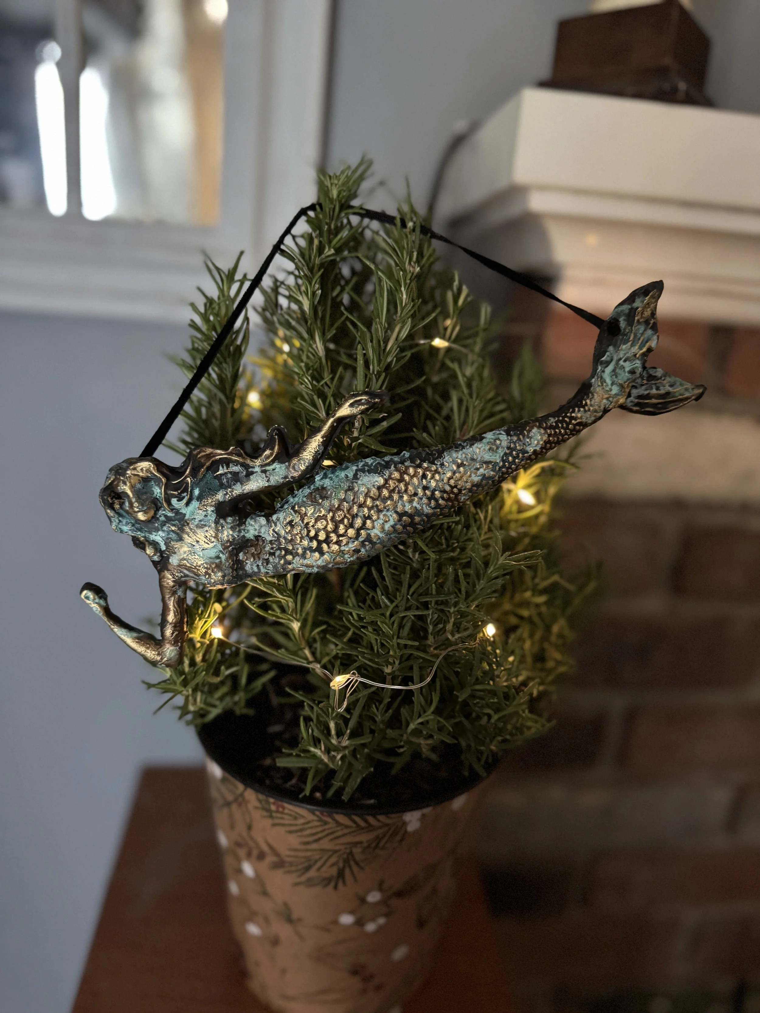 A small potted Christmas tree with a tree topper ornament shaped like a mermaid with its tail extended upward, green foliage, and string lights.