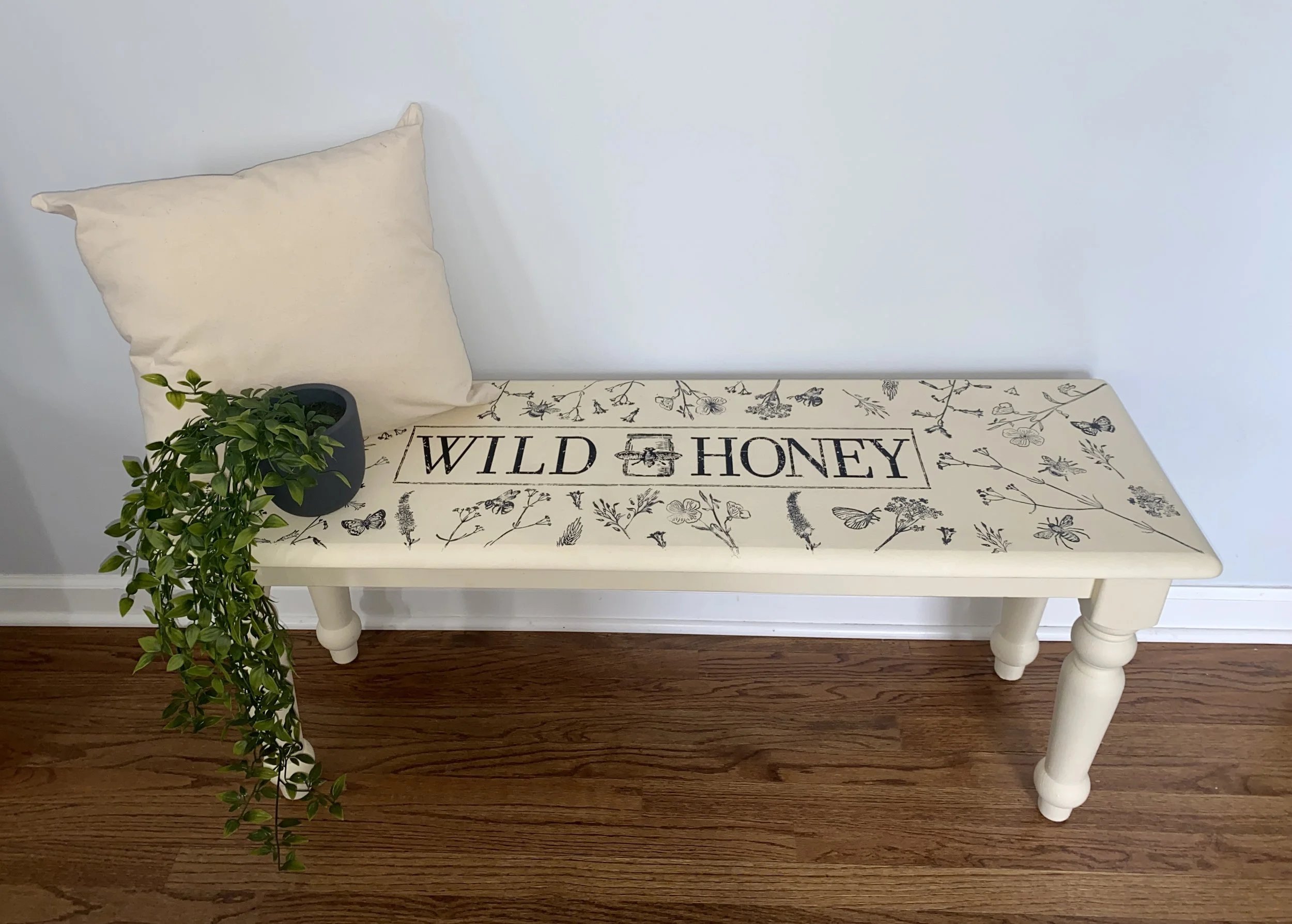 A white wooden bench with a beige pillow on the left side, a green potted plant on the left front corner, and a decorative fabric cover with black floral and insect designs that reads 'Wild Honey' in the center, set against a plain light blue wall on