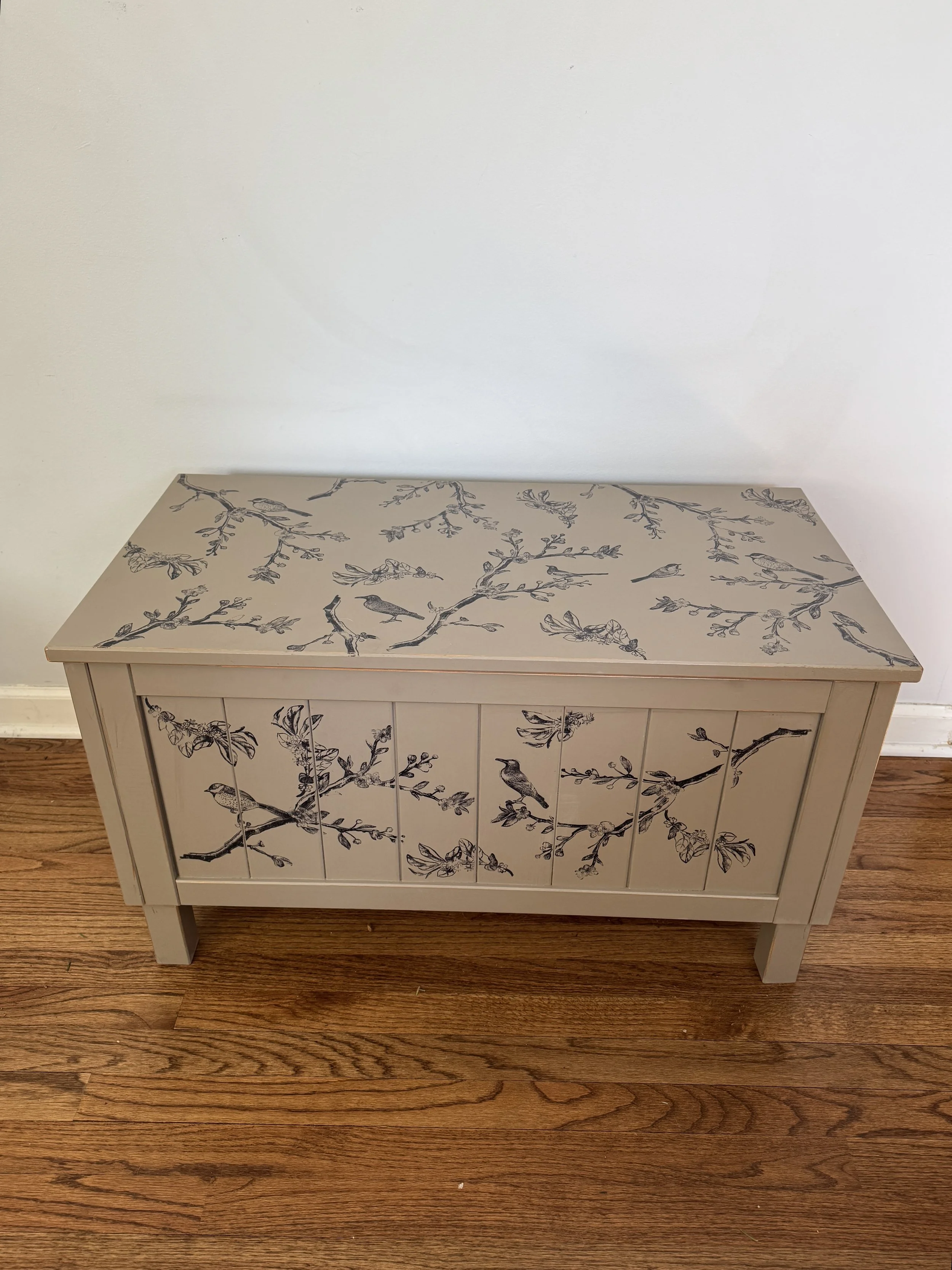 White wooden cabinet with floral and bird pattern on the front and top surface, placed on a hardwood floor against a plain wall.