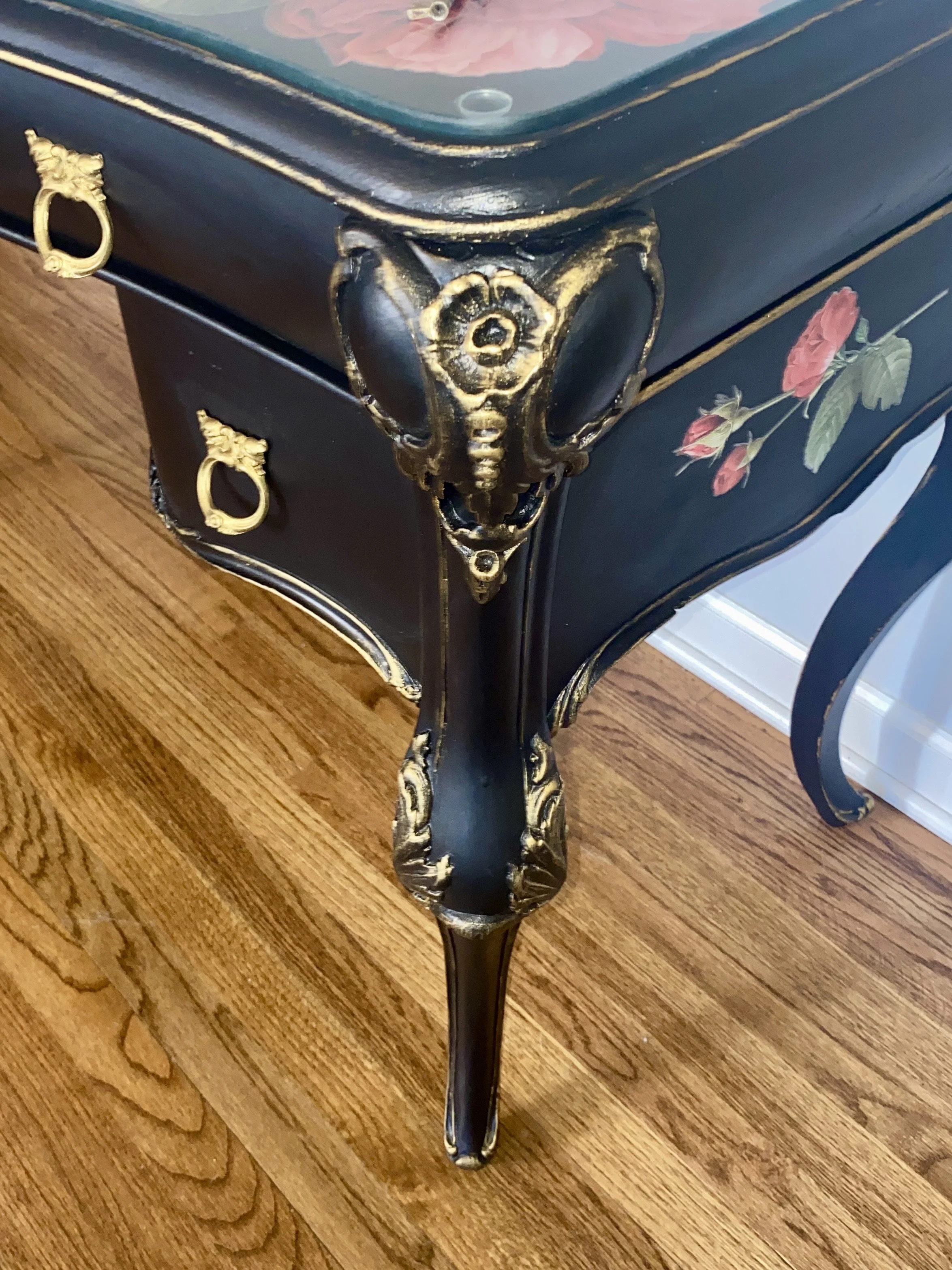 Close-up of a black vintage-style furniture piece with ornate gold accents, floral paintings, and curved legs, placed on a wooden floor.